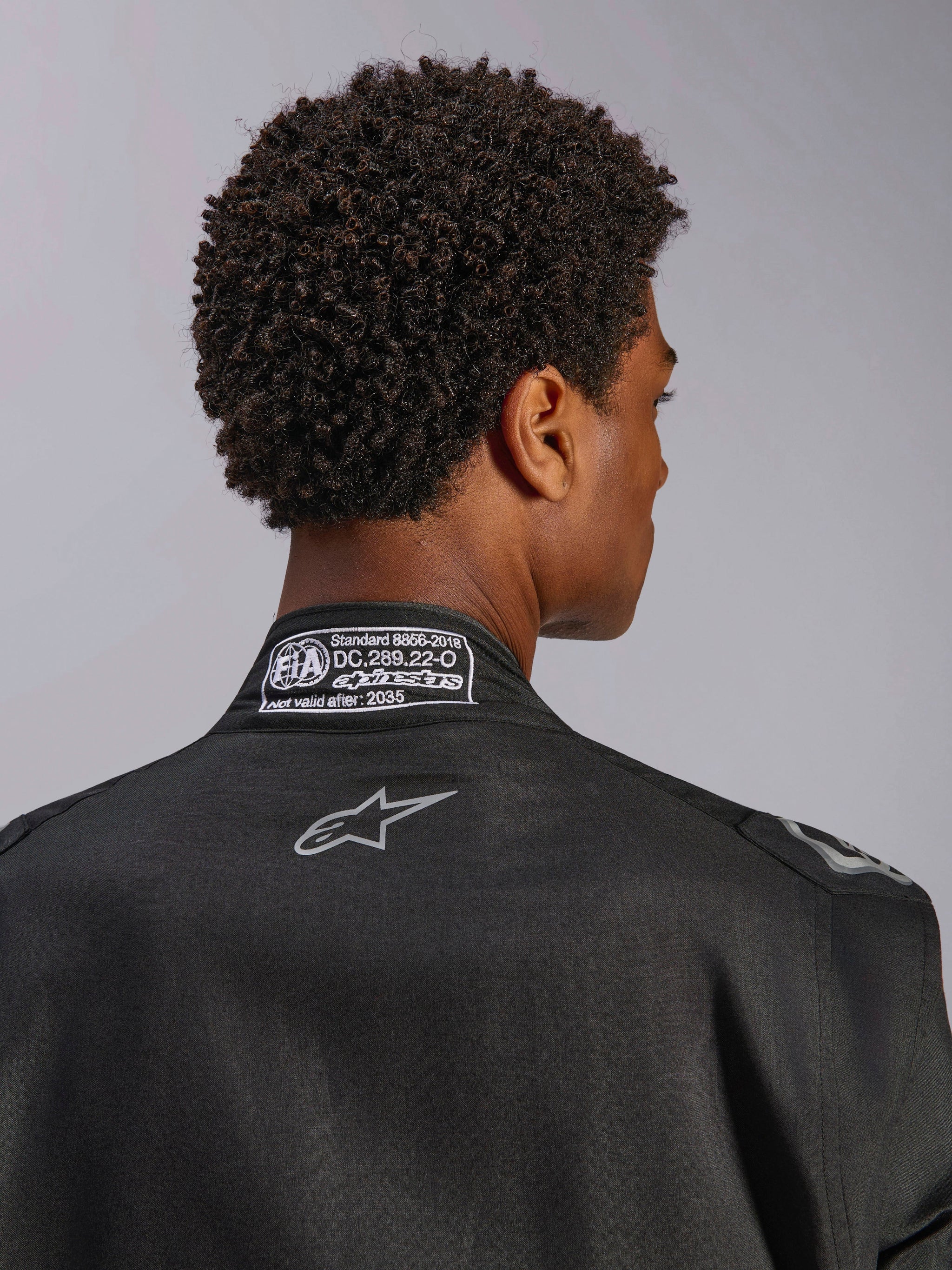 Alpinestars Hypertech V3 Suit, Black Dark Shadow, close-up rear view of the collar with FIA 8856-2018 certification label and back logo, professional lightweight racing suit with athletic fit