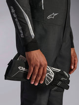Alpinestars Hypertech V3 Suit in Black Dark Shadow, close-up detail of the sleeve and torso with silver Alpinestars branding, hand holding a black racing glove with white accents, professional auto racing gear
