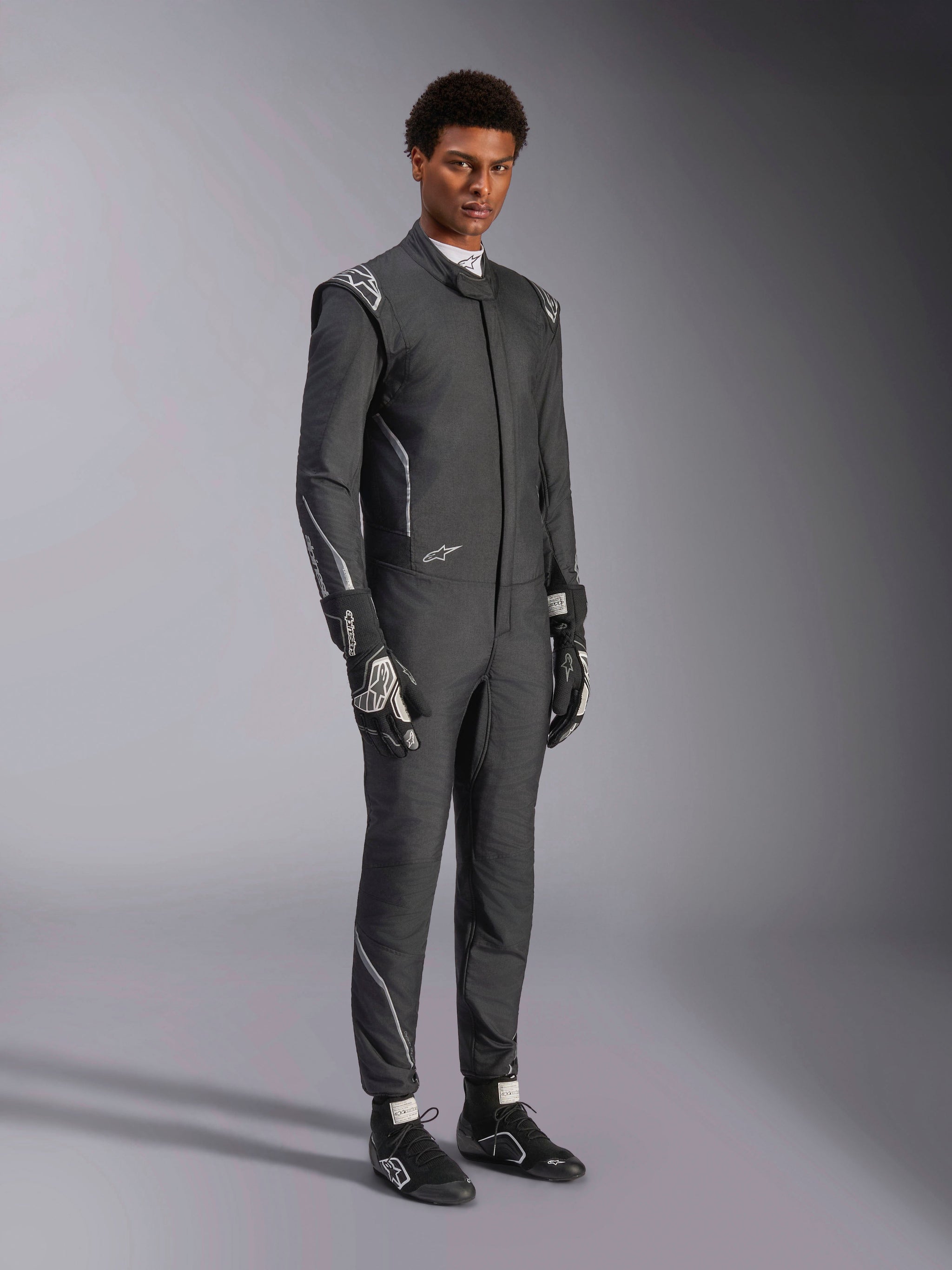Alpinestars Hypertech V3 Suit, Auto Racing Suit, Black and Dark Shadow, worn by male model with matching black gloves and racing shoes, athletic slim fit, features Alpinestars logos and light gray accents, professional motorsport protective gear