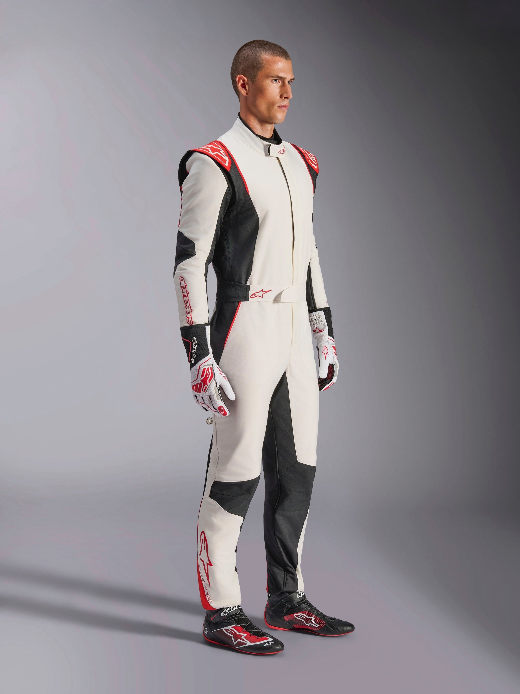 Male racing driver wearing Alpinestars GP Tech V4 Suit, White Black and Red, full-length one-piece auto racing suit, matching gloves and racing shoes, professional motorsport gear with Alpinestars branding and ergonomic design
