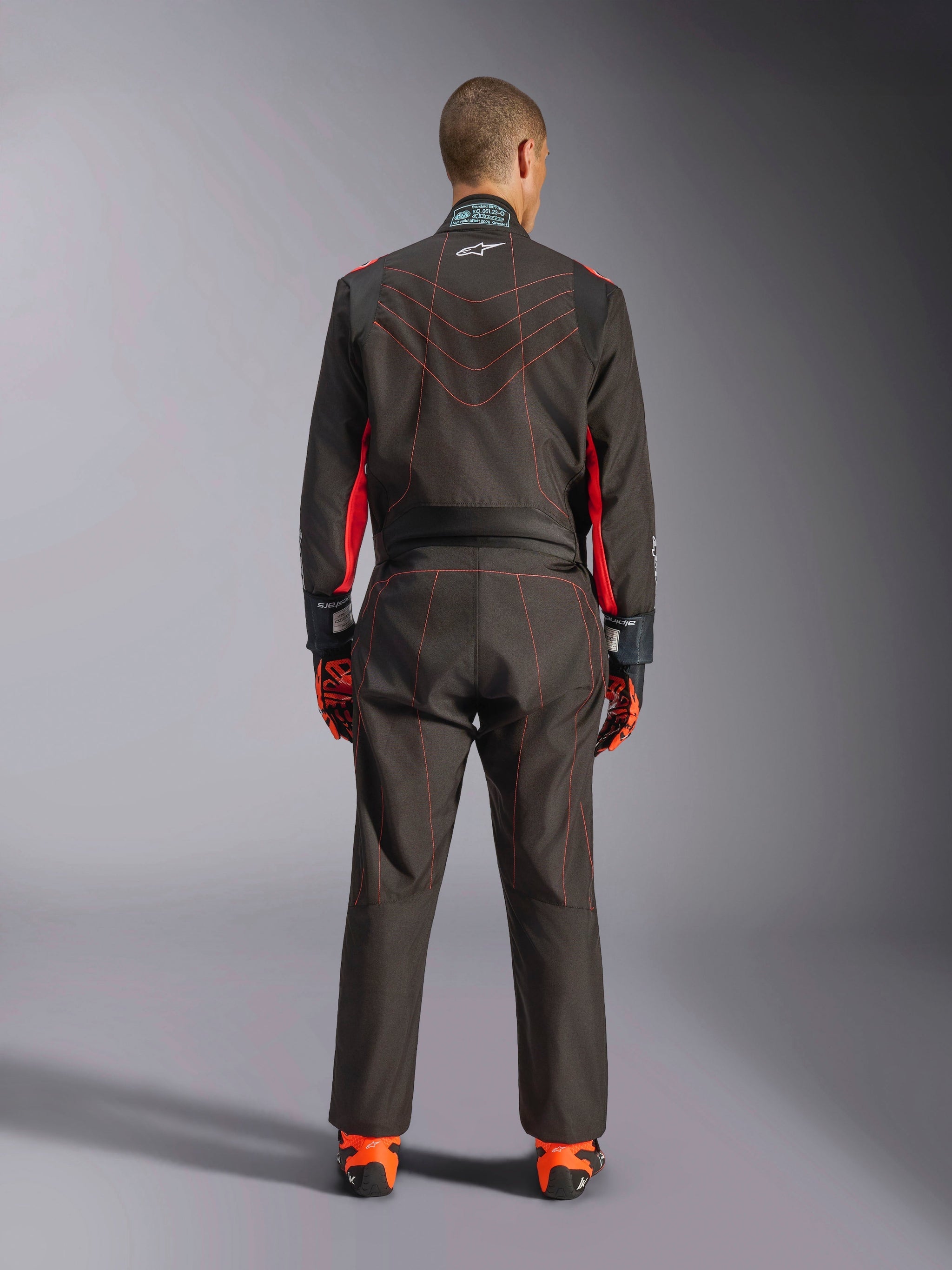 Rear view of a driver wearing the Alpinestars KMX-9 V3 Suit, Karting Suit, Black with Red Fluo accents and red contrast stitching, professional motorsport racing gear, worn with orange and black racing gloves and shoes.