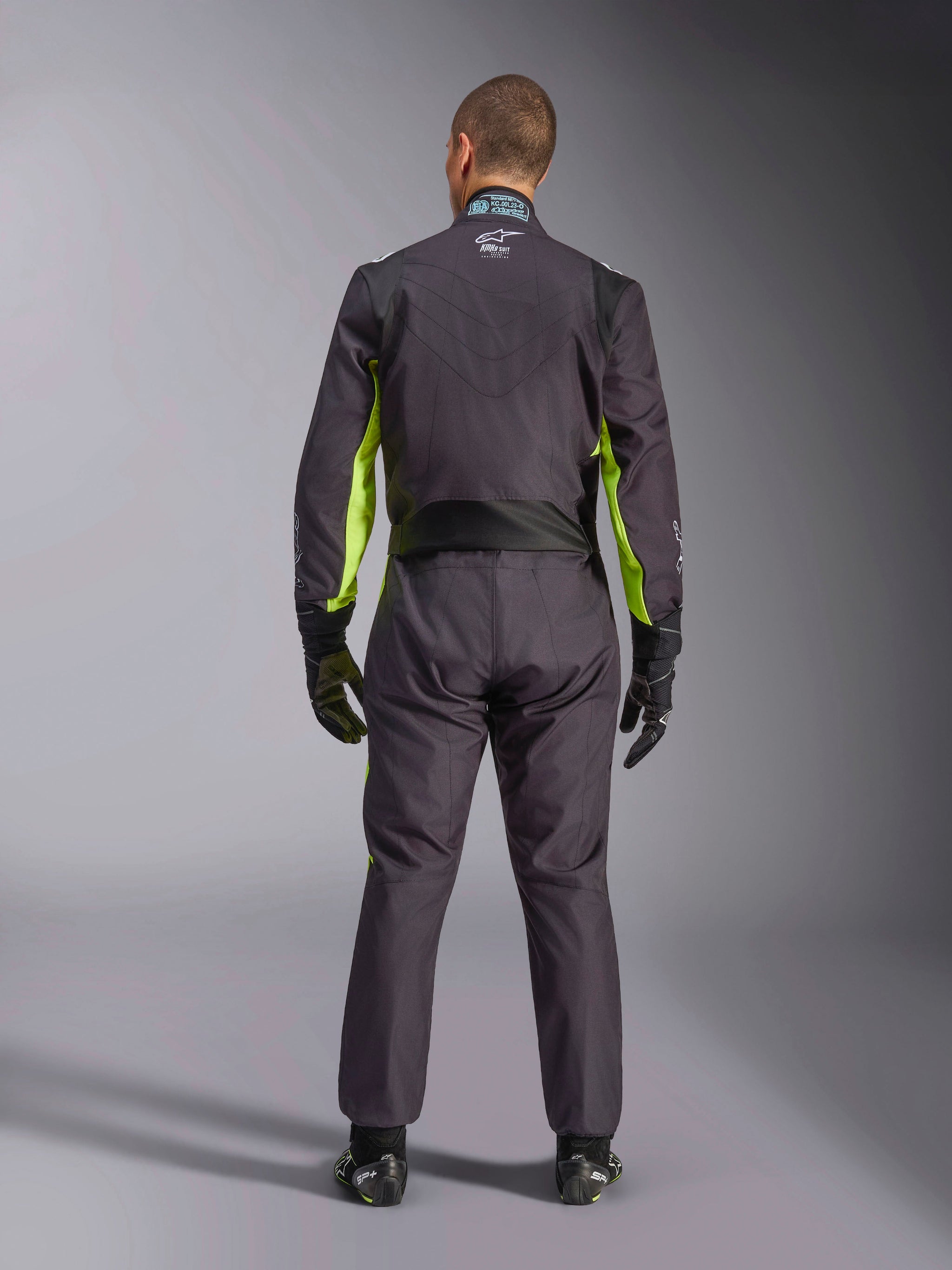Full-body rear view of a person wearing the Alpinestars KMX-9 V3 Graphic 3 Suit, Karting Suit in Black and Yellow Fluo, showing ergonomic stretch panels on the back and shoulders, paired with black racing gloves and footwear.