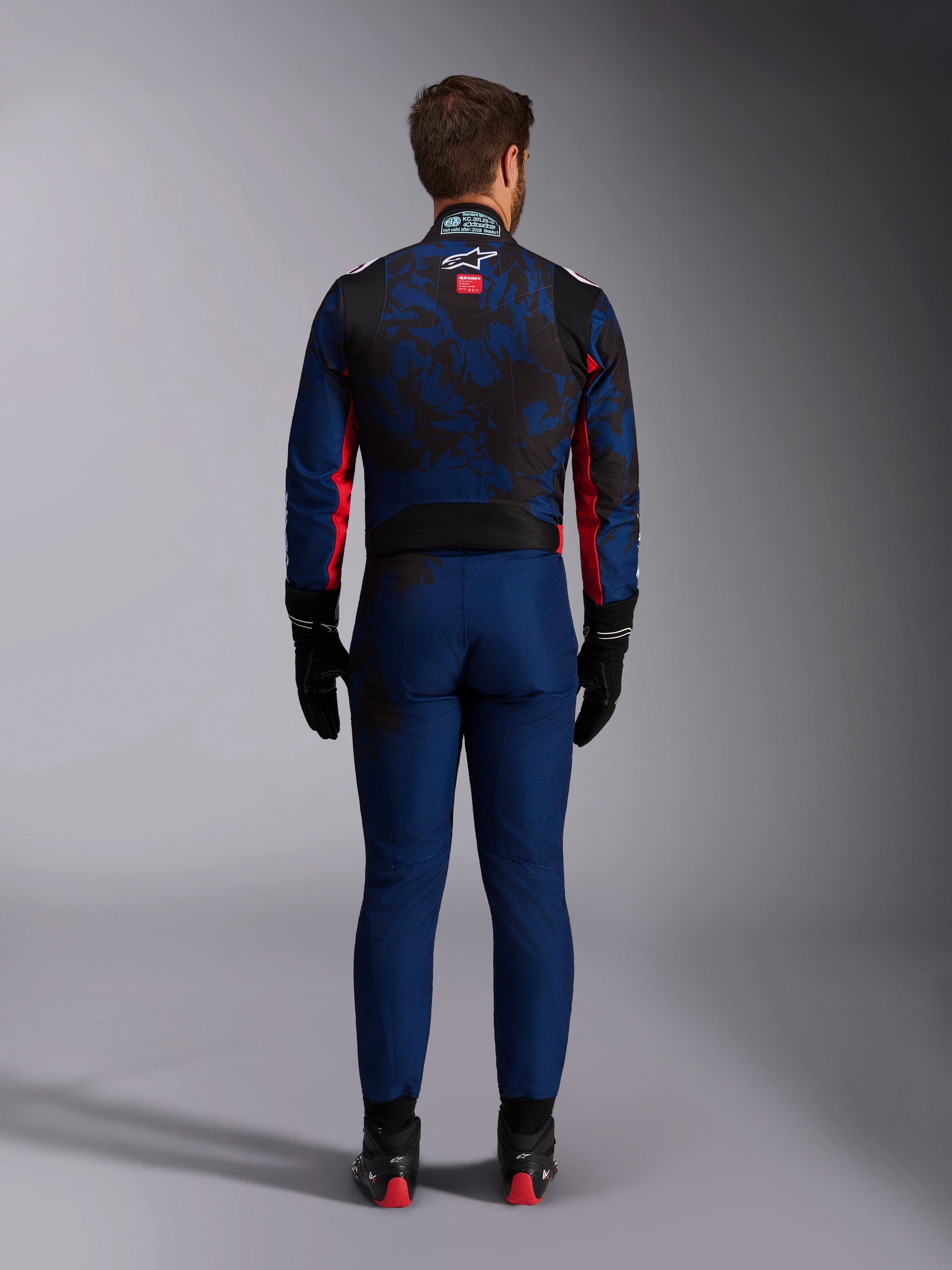Karting driver from the back wearing the KMX-9 V3 Graphic 5 Suit in Navy Red, featuring a navy blue and black graphic pattern, red side accents, and the Alpinestars logo on the upper back, paired with black racing gloves and footwear.