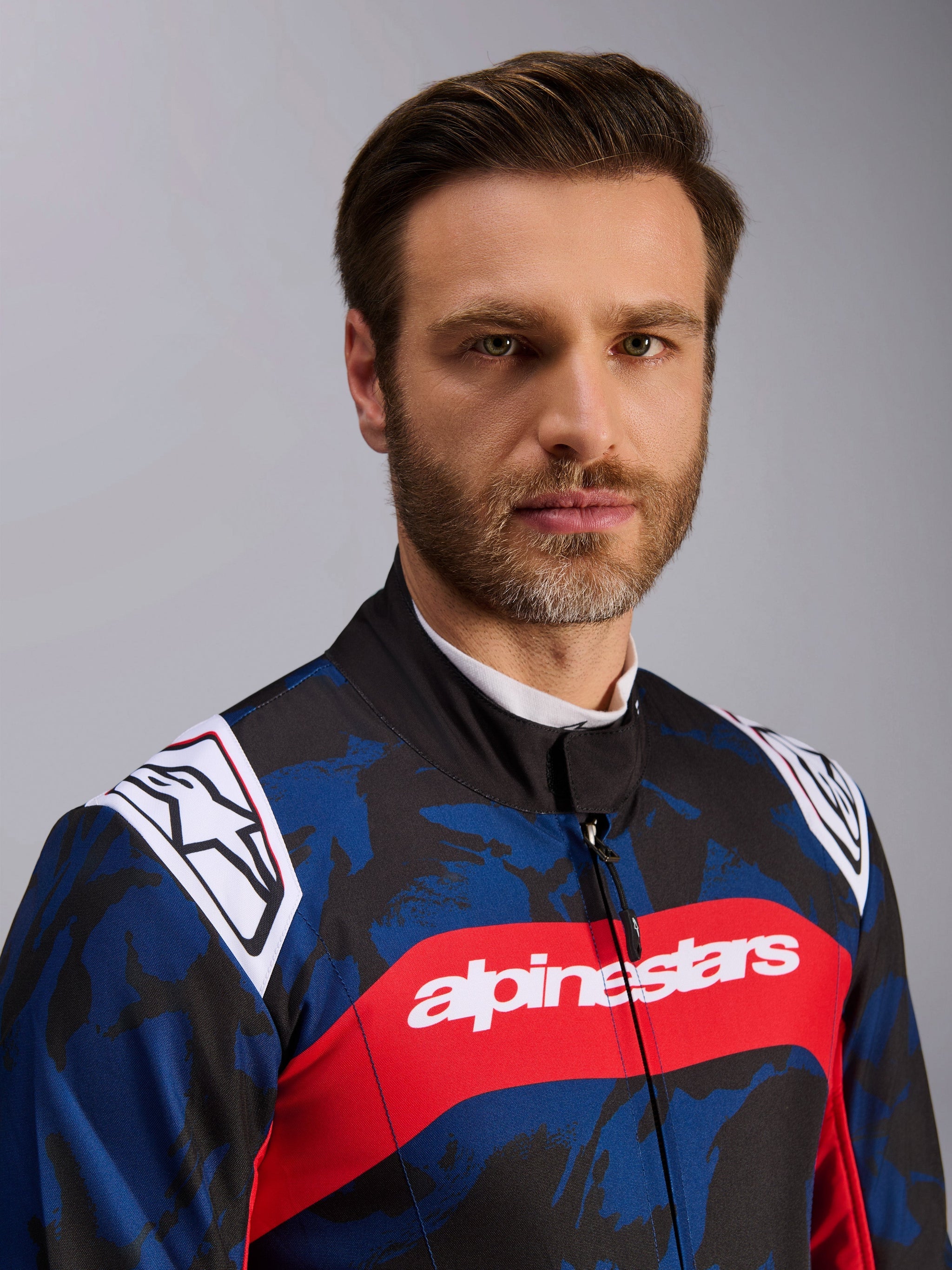 Alpinestars KMX-9 V3 Graphic 5 Suit, Karting Suit in Navy Red, featuring a blue and black camouflage graphic pattern, bold red horizontal stripe with white alpinestars text across the chest, and white logos on the shoulders, worn by a model in a studio setting.