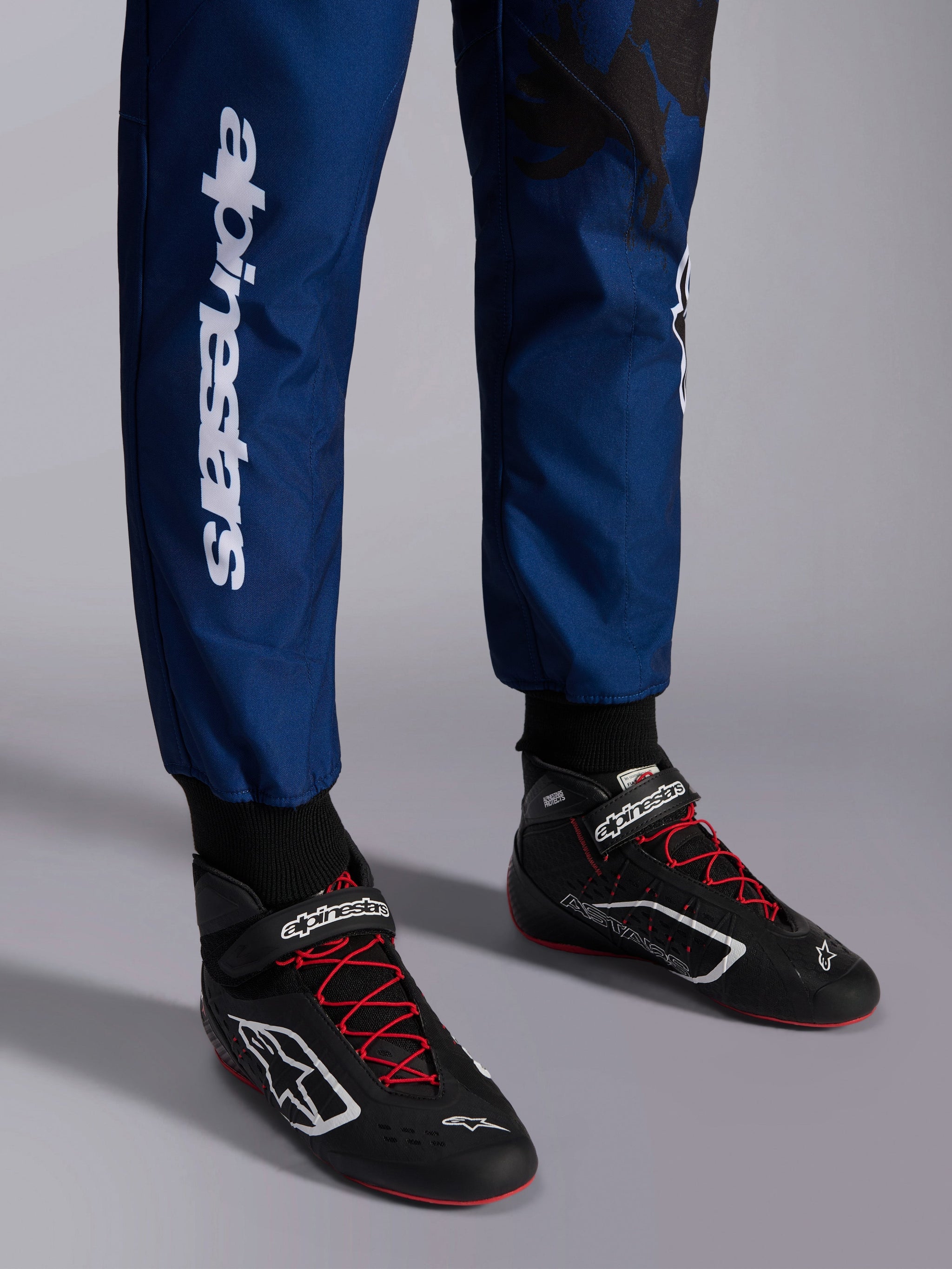 Alpinestars KMX-9 V3 Graphic 5 Suit, Navy Red, close-up of lower legs and karting shoes, navy blue fabric with white vertical Alpinestars branding, black shoes with red laces and white logos, professional karting gear