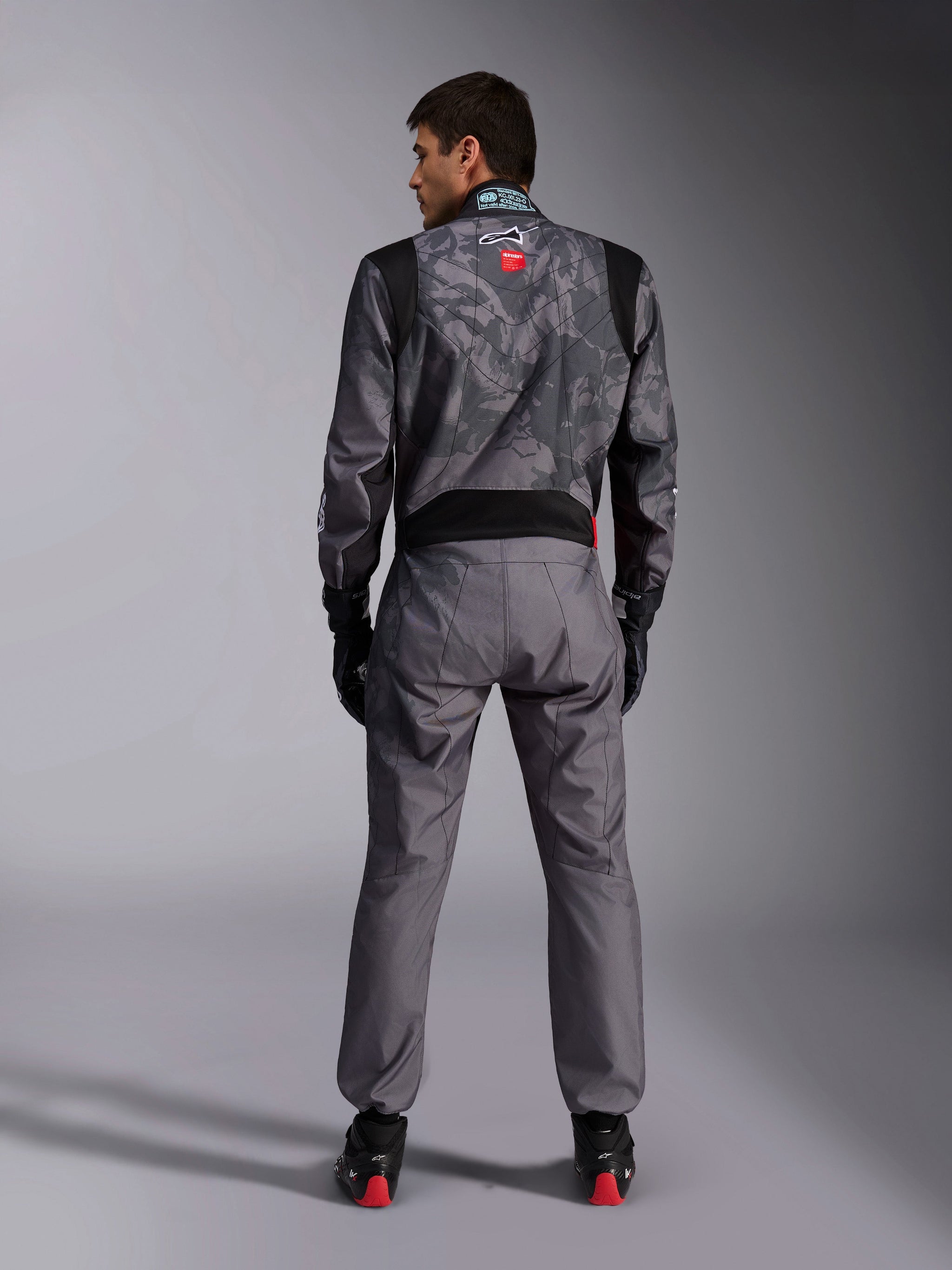 Alpinestars KMX-9 V3 Graphic 5 Suit, Karting Suit, Dark Gray and Black camouflage pattern on the upper body, back view showing stretch panels and Alpinestars branding, worn with black gloves and black racing shoes with red details, professional motorsport racing gear