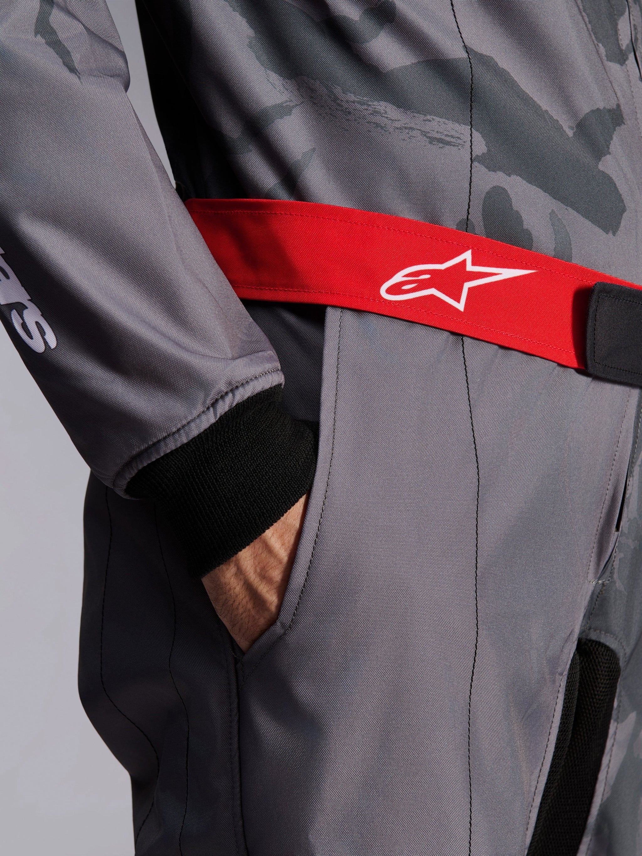 Alpinestars KMX-9 V3 Graphic 5 Suit, Karting Suit, Dark Gray and Black with Red belt, close-up featuring red adjustable waist belt with white logo, hand in thigh pocket, and black knitted cuff on gray graphic patterned fabric