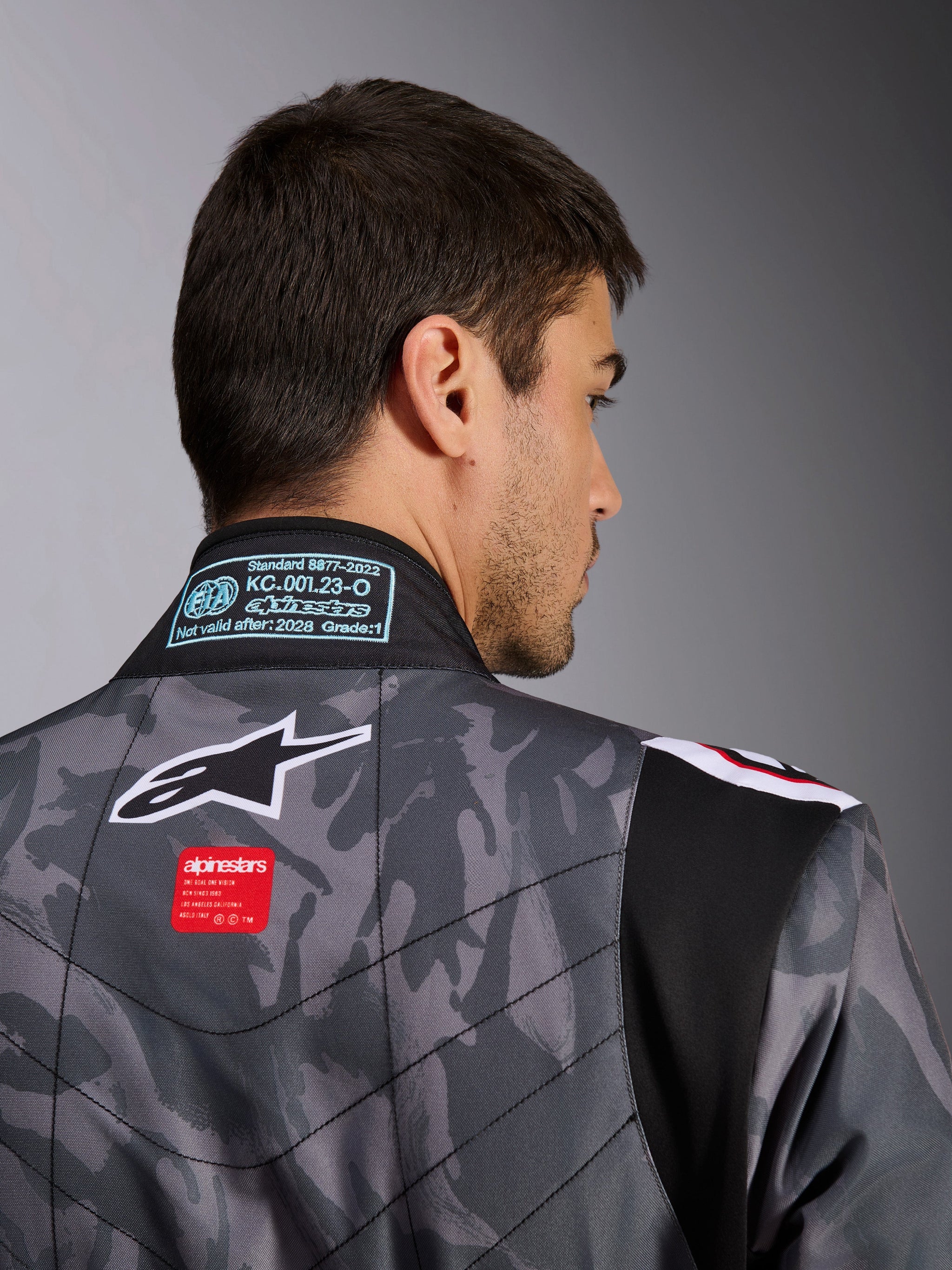 Alpinestars KMX-9 V3 Graphic 5 Suit, Karting Suit, Dark Gray and Black camouflage pattern, close-up of the upper back showing the Alpinestars logo, red branding tag, and FIA homologation label on the collar.