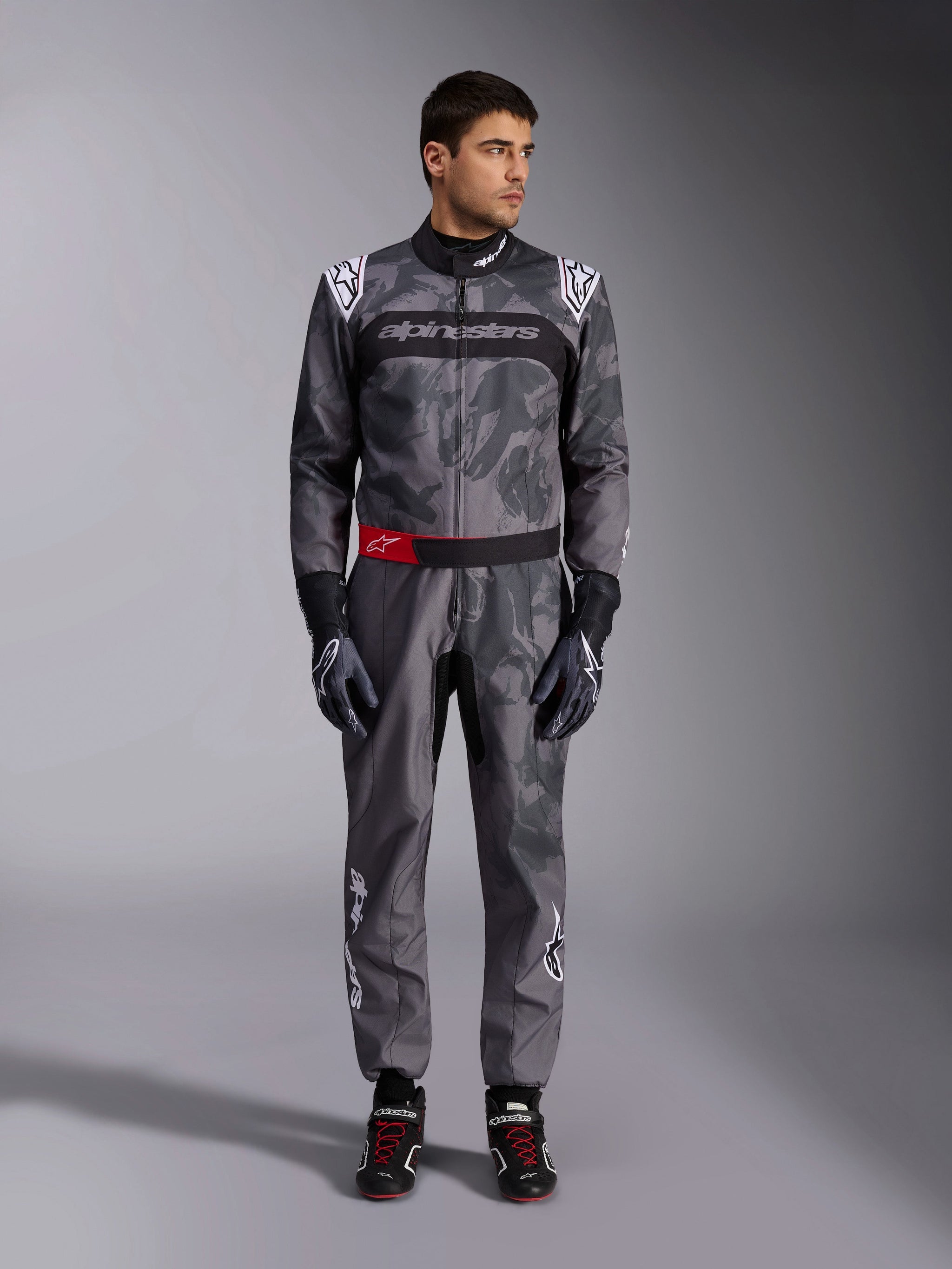 Alpinestars KMX-9 V3 Graphic 5 Suit, Karting Suit, Dark Gray and Black with camouflage-style graphics, featuring a black chest band with white logo and a red waist belt accent, worn with matching racing gloves and shoes, professional motorsport gear
