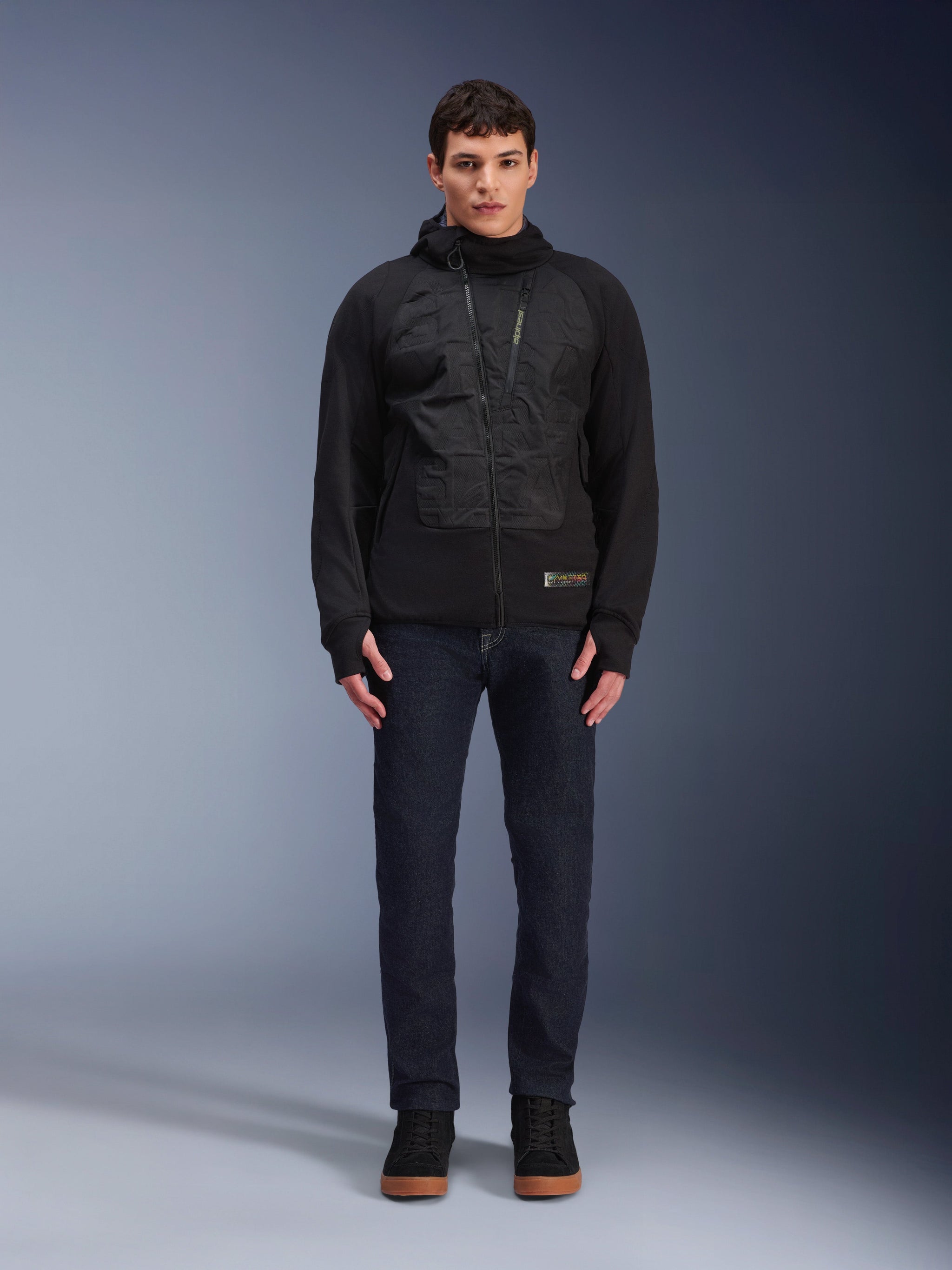 Alpinestars MO.ST.EQ Hybrid Hooded Jacket, Black urban motorcycle jacket worn by a model with dark jeans and black high-top sneakers, featuring an angled front zipper and padded chest panels, modern streetwear-inspired riding gear
