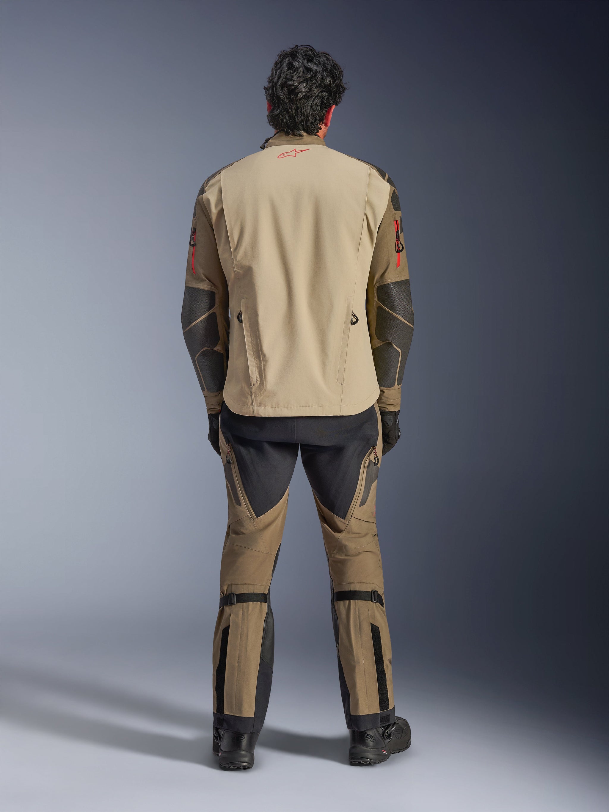 Alpinestars Nazca 3L GORE-TEX PRO Jacket in Walnut Rock Brown, back view of rider, showing Superfabric reinforcements on elbows and rear ventilation zippers, worn with matching adventure pants, black boots, and gloves, rugged technical riding apparel for extreme adventure.