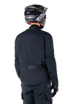 Alpinestars St-7 Prime GORE-TEX Jacket, Black, rear view of a rider wearing the jacket with a matching helmet and pants, featuring Alpinestars branding and geographic coordinates on the lower back.