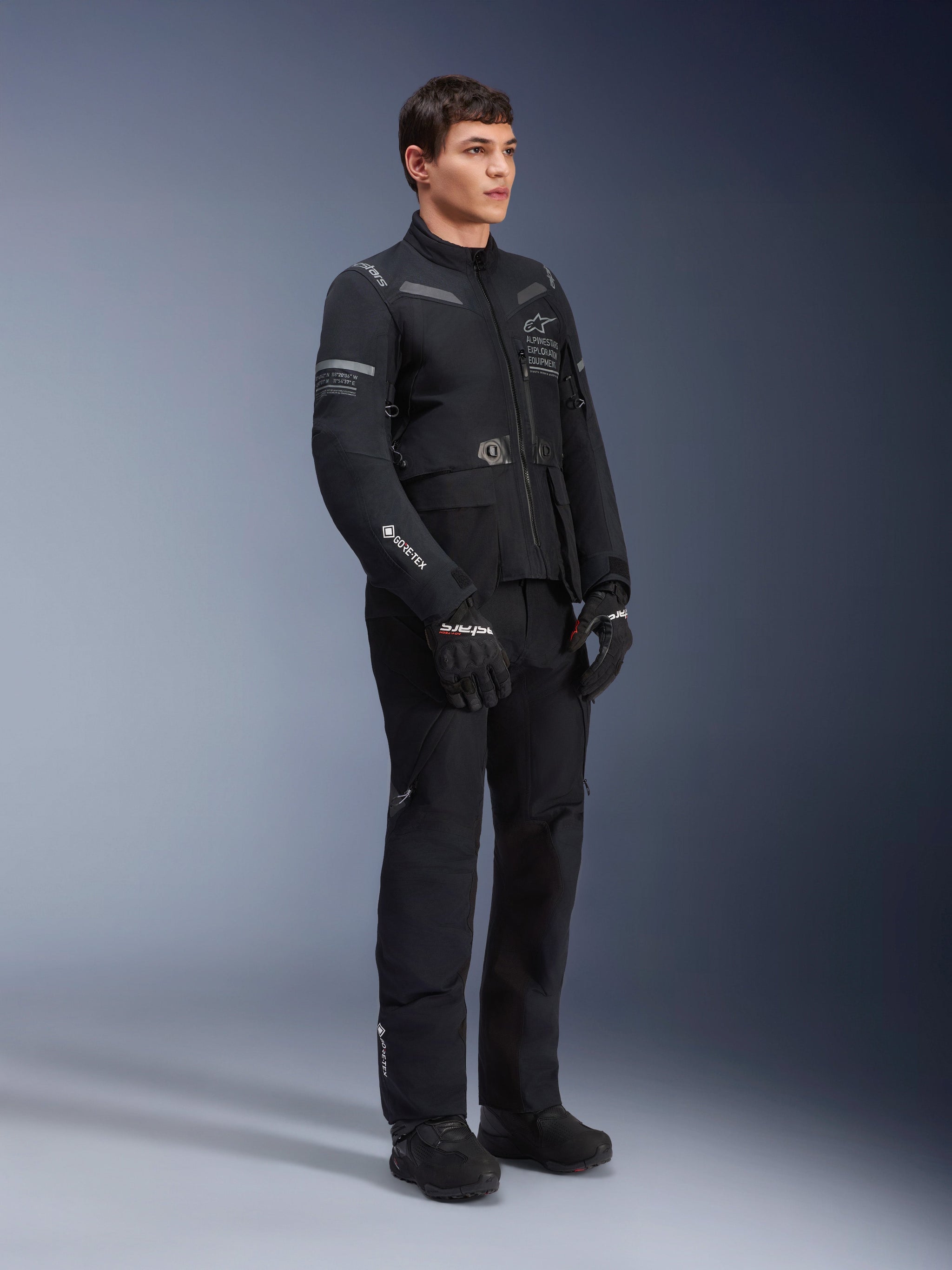 Alpinestars ST-7 2L GORE-TEX Pants, worn by a model with the matching ST-7 Jacket, all-black adventure touring motorcycle outfit, featuring GORE-TEX branding, cargo pockets, and black gloves and boots, studio shot against a gray background