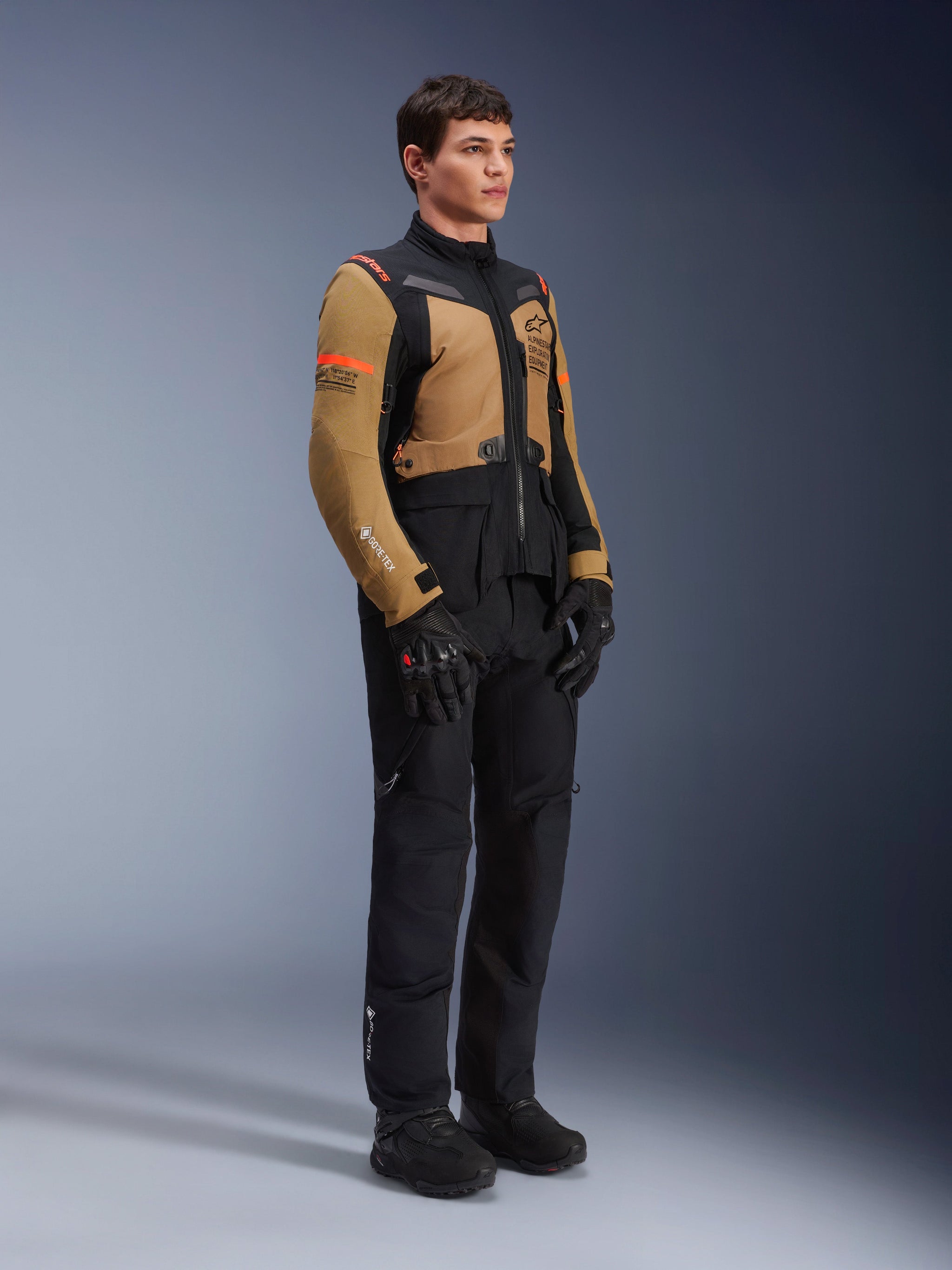Alpinestars ST-7 2L GORE-TEX Jacket, Motorcycle Jacket, Gobi Brown and Black, worn by a model with matching black pants and gloves, featuring brown panels on the chest and sleeves, black technical accents, and orange branding, adventure-touring motorcycle gear.