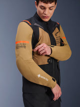 Alpinestars ST-7 2L GORE-TEX Jacket in Gobi Brown and Black, adventure-touring motorcycle jacket worn by a model adjusting a sleeve vent, featuring orange accents, coordinate graphics, and GORE-TEX logo