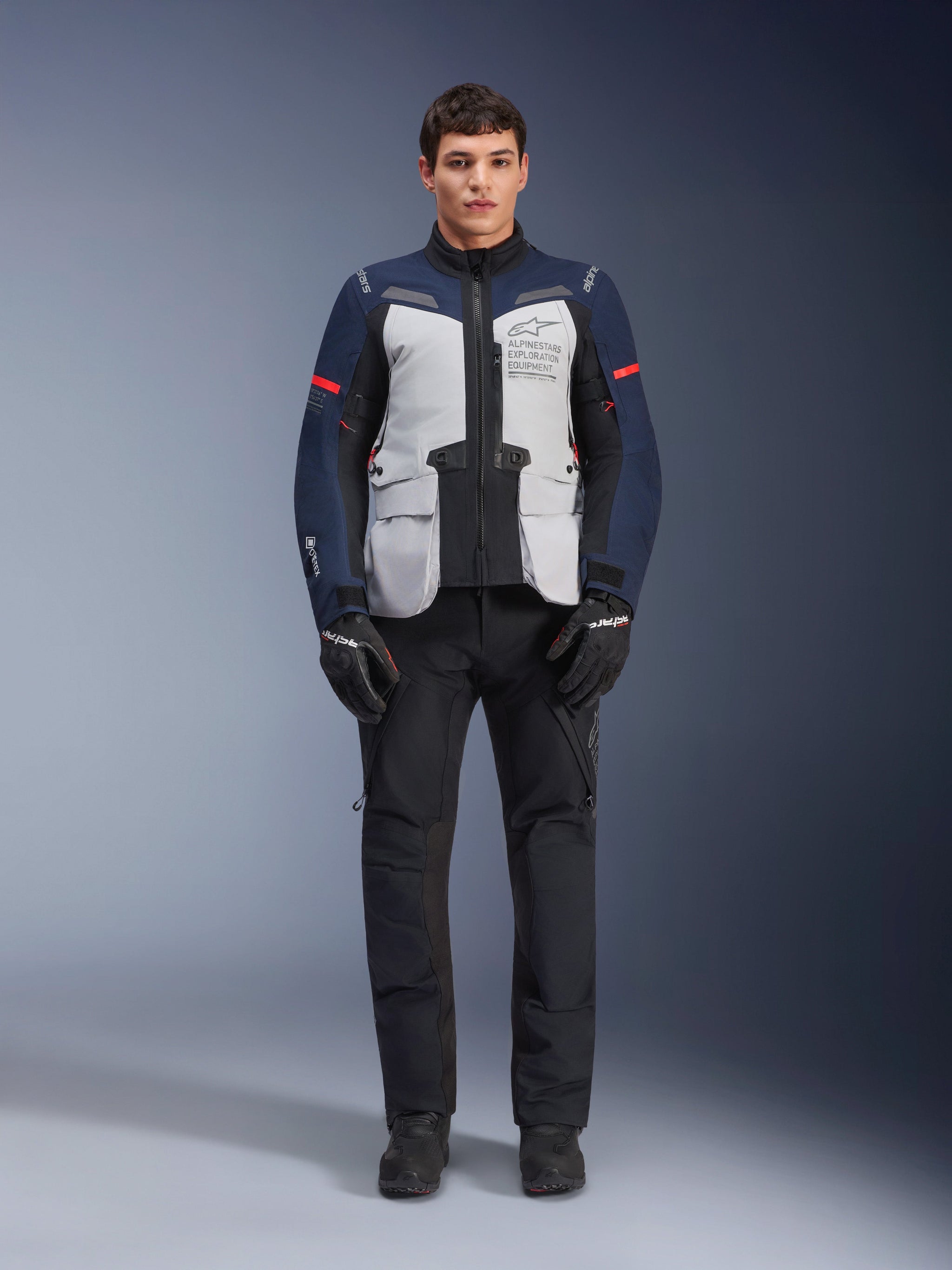 Alpinestars ST-7 2L GORE-TEX Jacket, Adventure Touring Motorcycle Jacket, Ice Gray Dark Blue and Black, worn with black pants and gloves, featuring large front cargo pockets, red accents on the sleeves, and a laminated GORE-TEX shell for waterproof performance.