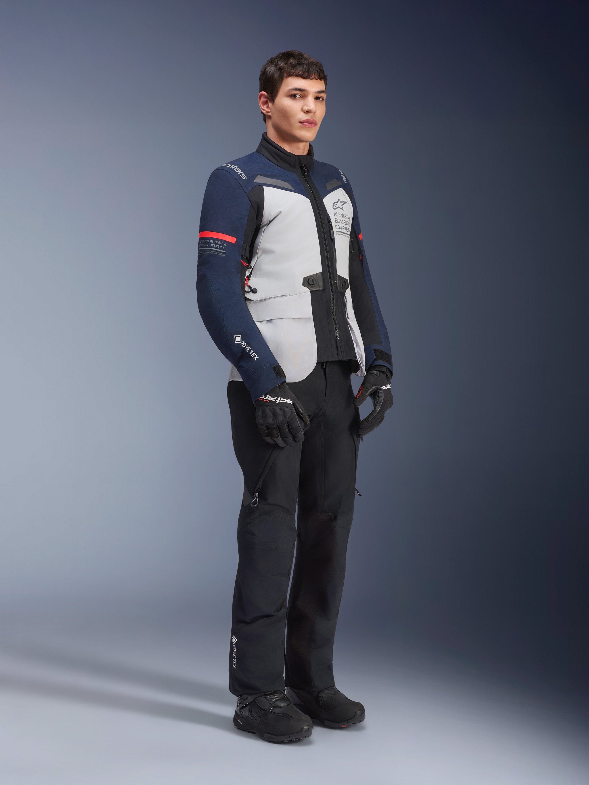 Alpinestars ST-7 2L GORE-TEX Jacket in Ice Gray, Dark Blue, and Black colorway with red accents, worn by a male model with black motorcycle pants and gloves, professional adventure touring motorcycle gear