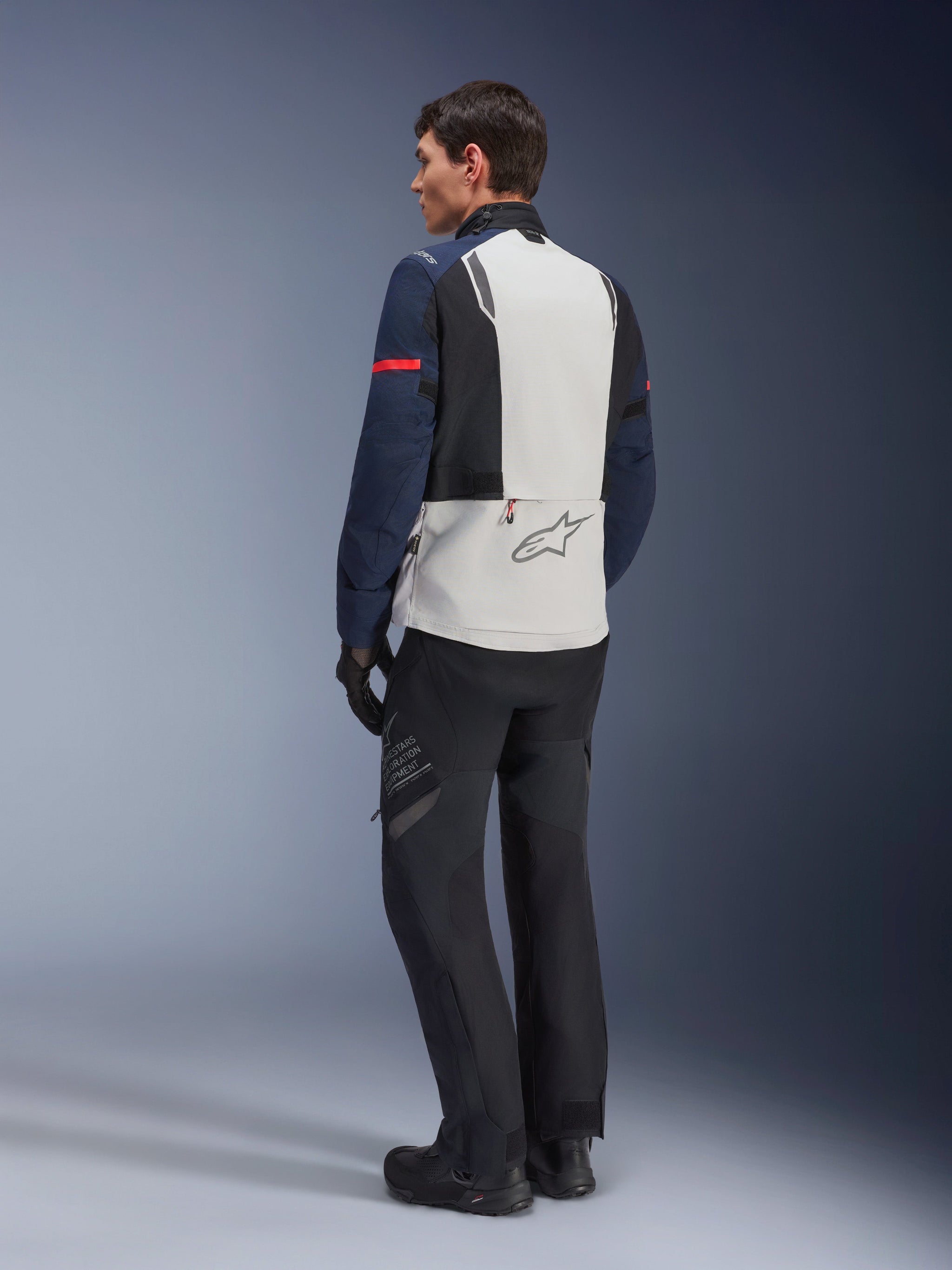 Model wearing Alpinestars ST-7 2L GORE-TEX Jacket in Ice Gray Dark Blue and Black, rear view, light gray back panel with Alpinestars logo, dark blue sleeves with red stripe, paired with black adventure riding pants and boots, motorcycle touring gear