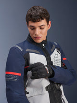 Alpinestars ST-7 2L GORE-TEX Jacket, Ice Gray Dark Blue and Black with orange accents, worn by a man adjusting a sleeve detail, featuring 'Alpinestars Exploration Equipment' branding and geographic coordinates, professional adventure motorcycle riding gear