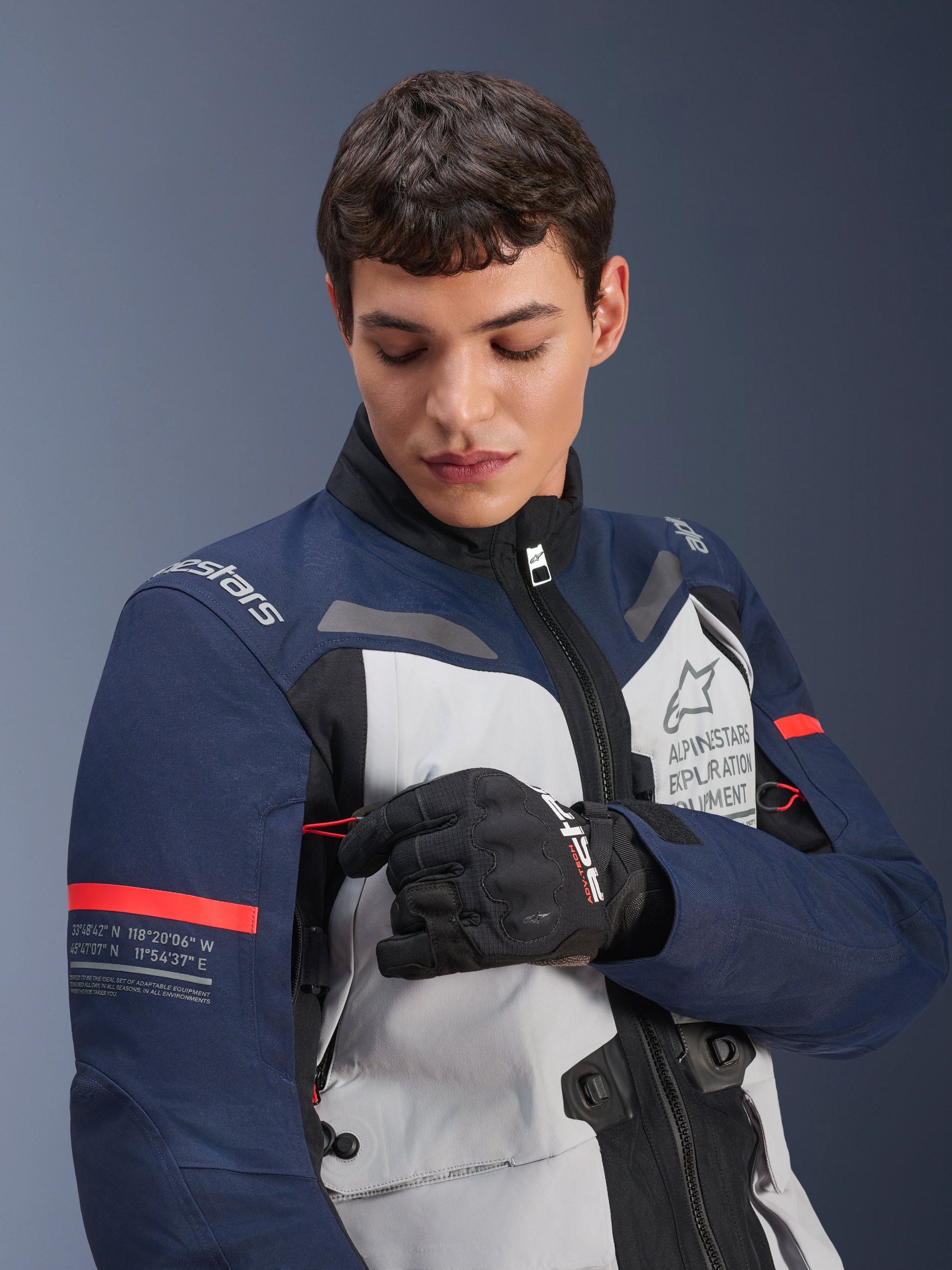Alpinestars ST-7 2L GORE-TEX Jacket, Ice Gray Dark Blue and Black with orange accents, worn by a man adjusting a sleeve detail, featuring 'Alpinestars Exploration Equipment' branding and geographic coordinates, professional adventure motorcycle riding gear