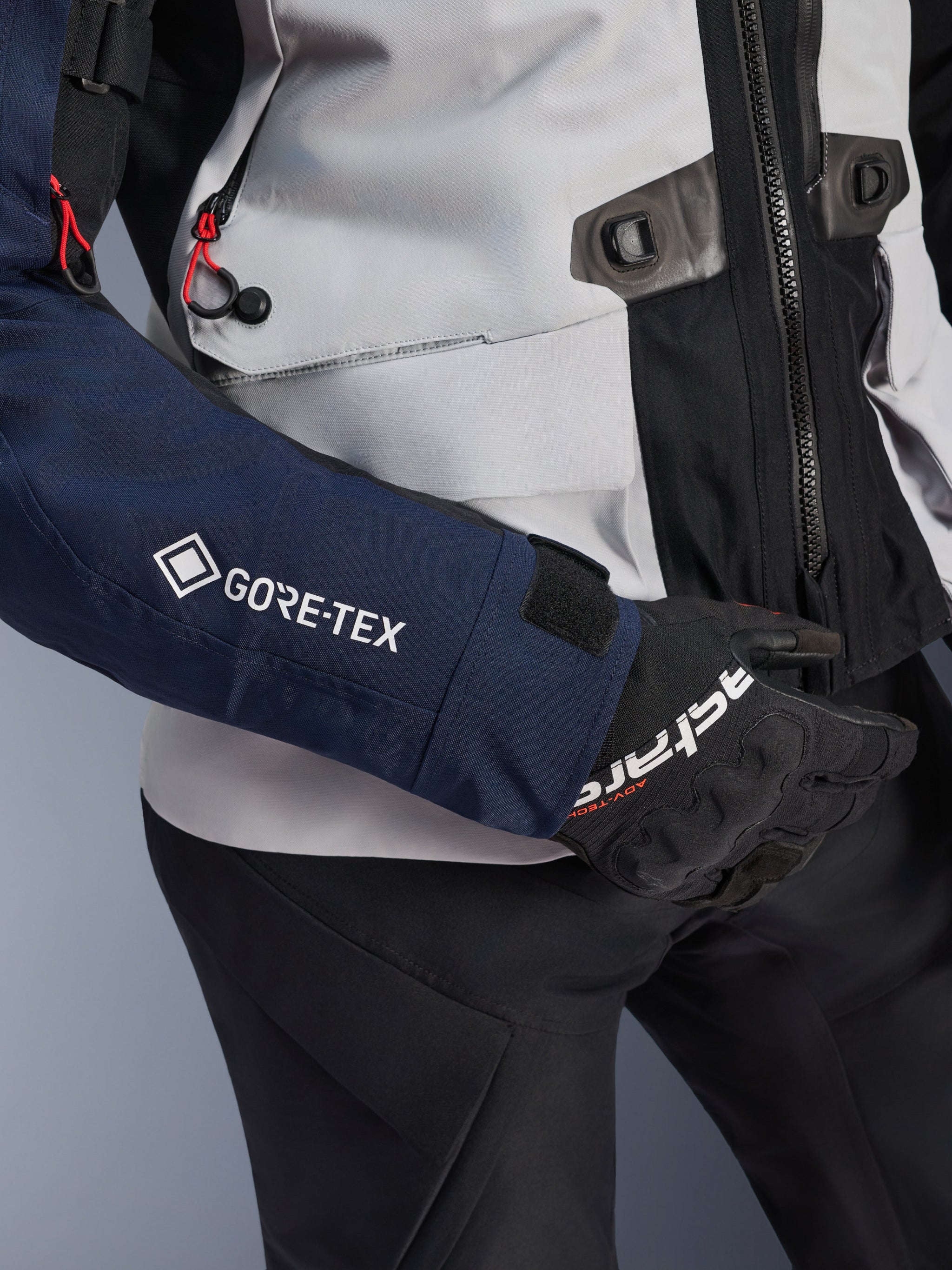 Alpinestars ST-7 2L GORE-TEX Jacket, close-up of dark blue sleeve with white GORE-TEX logo, ice gray torso with black detailing and red cord accents, worn with black Alpinestars gloves and pants, adventure touring motorcycle gear