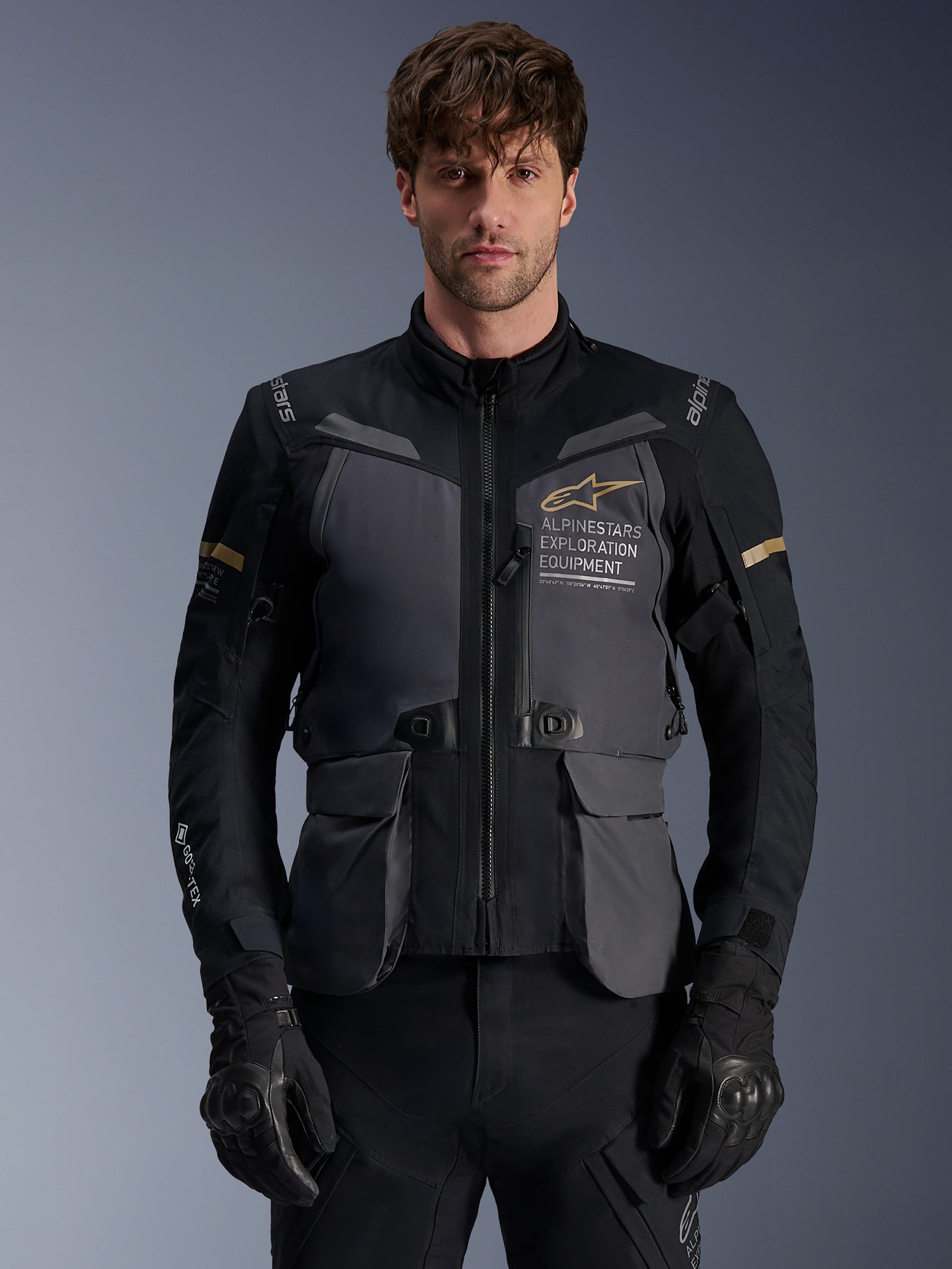 Alpinestars ST-7 2L GORE-TEX Jacket, Men's Adventure Touring Motorcycle Jacket, Deep Gray and Black with Khaki accents, featuring 'Alpinestars Exploration Equipment' branding, waterproof cargo pockets, and high collar, worn by a model with black gloves