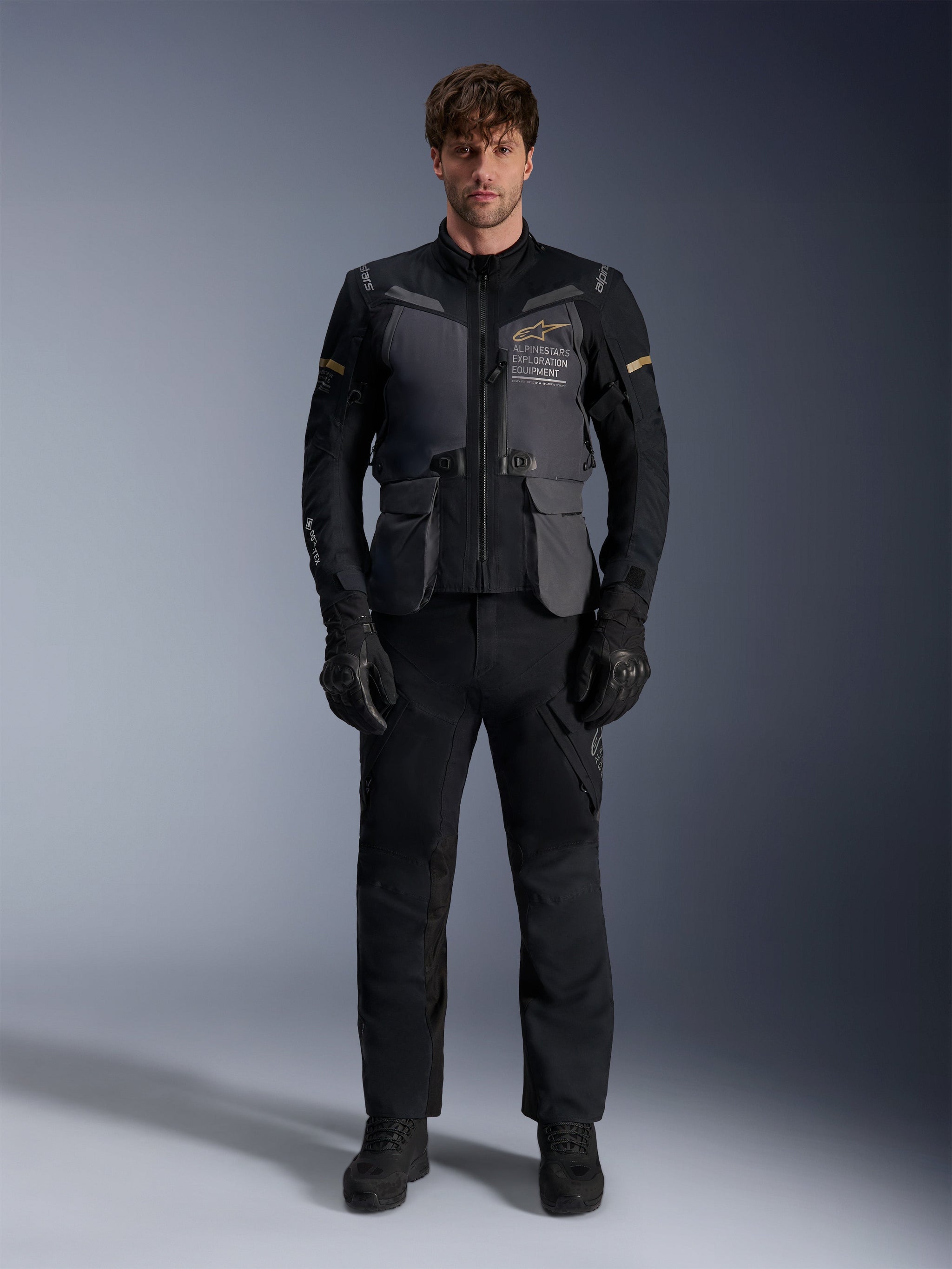 Male model wearing the Alpinestars ST-7 2L GORE-TEX Jacket in Deep Gray Black Dark Khaki, featuring 'ALPINESTARS EXPLORATION EQUIPMENT' branding on the chest, paired with matching black riding pants, gloves, and boots, studio front view.