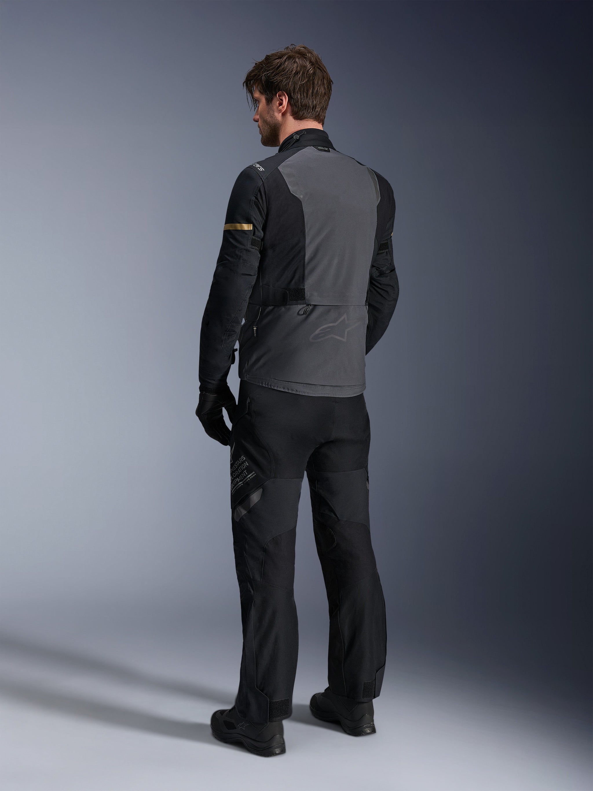 Alpinestars ST-7 2L GORE-TEX Jacket, Deep Gray and Black with Dark Khaki accents, rear view of a rider wearing the jacket and matching motorcycle pants, adventure touring gear with waterproof laminated construction