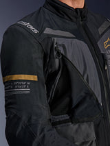 Alpinestars ST-7 2L GORE-TEX Jacket, Deep Gray and Black, close-up showing open zippered chest ventilation panel with mesh, Alpinestars logo on shoulder, and khaki stripe on the sleeve with printed geographic coordinates, technical adventure-touring motorcycle gear
