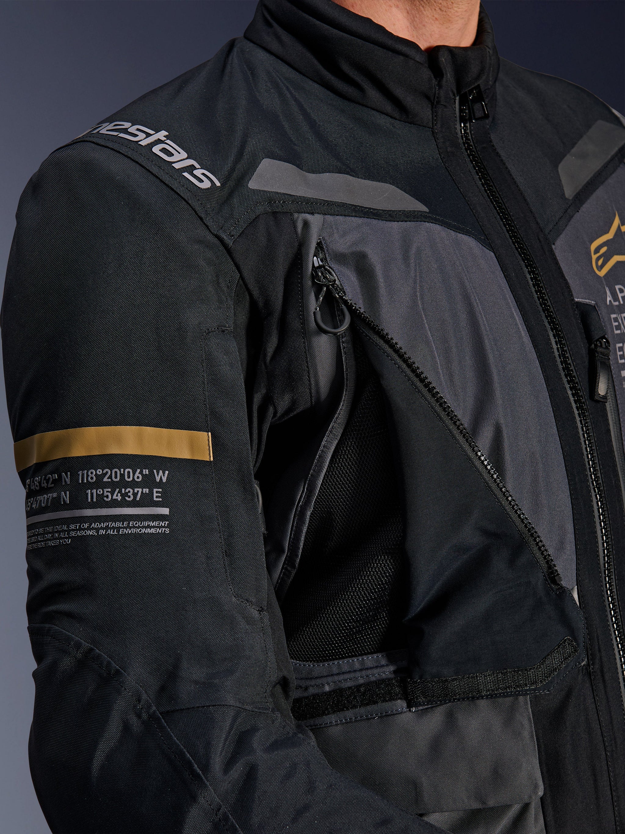Alpinestars ST-7 2L GORE-TEX Jacket, Deep Gray and Black, close-up showing open zippered chest ventilation panel with mesh, Alpinestars logo on shoulder, and khaki stripe on the sleeve with printed geographic coordinates, technical adventure-touring motorcycle gear