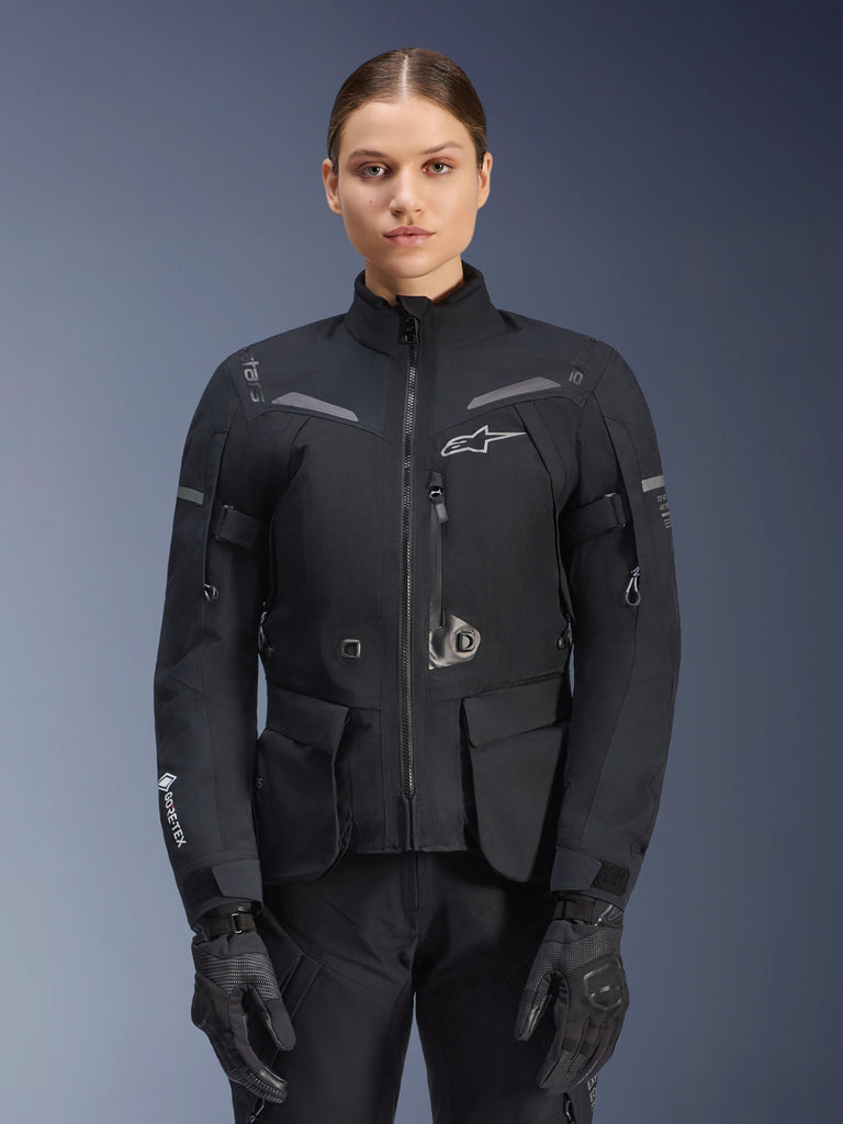 Woman wearing Alpinestars Stella ST-7 2L GORE-TEX Jacket, Black with Dark Gray accents, motorcycle touring jacket with large cargo pockets and high collar, paired with black gloves and pants, waterproof adventure riding apparel