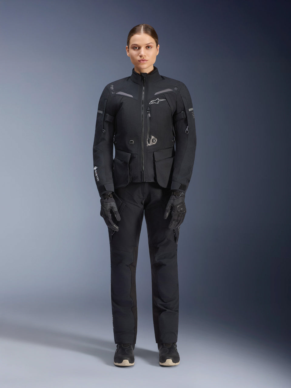 Female rider wearing Alpinestars Women Stella ST-7 2L GORE-TEX Jacket and Pants, Black motorcycle touring outfit, waterproof and breathable technical gear, worn with matching black gloves and boots, full-body protective gear with reflective elements, modern technical design for adventure riding