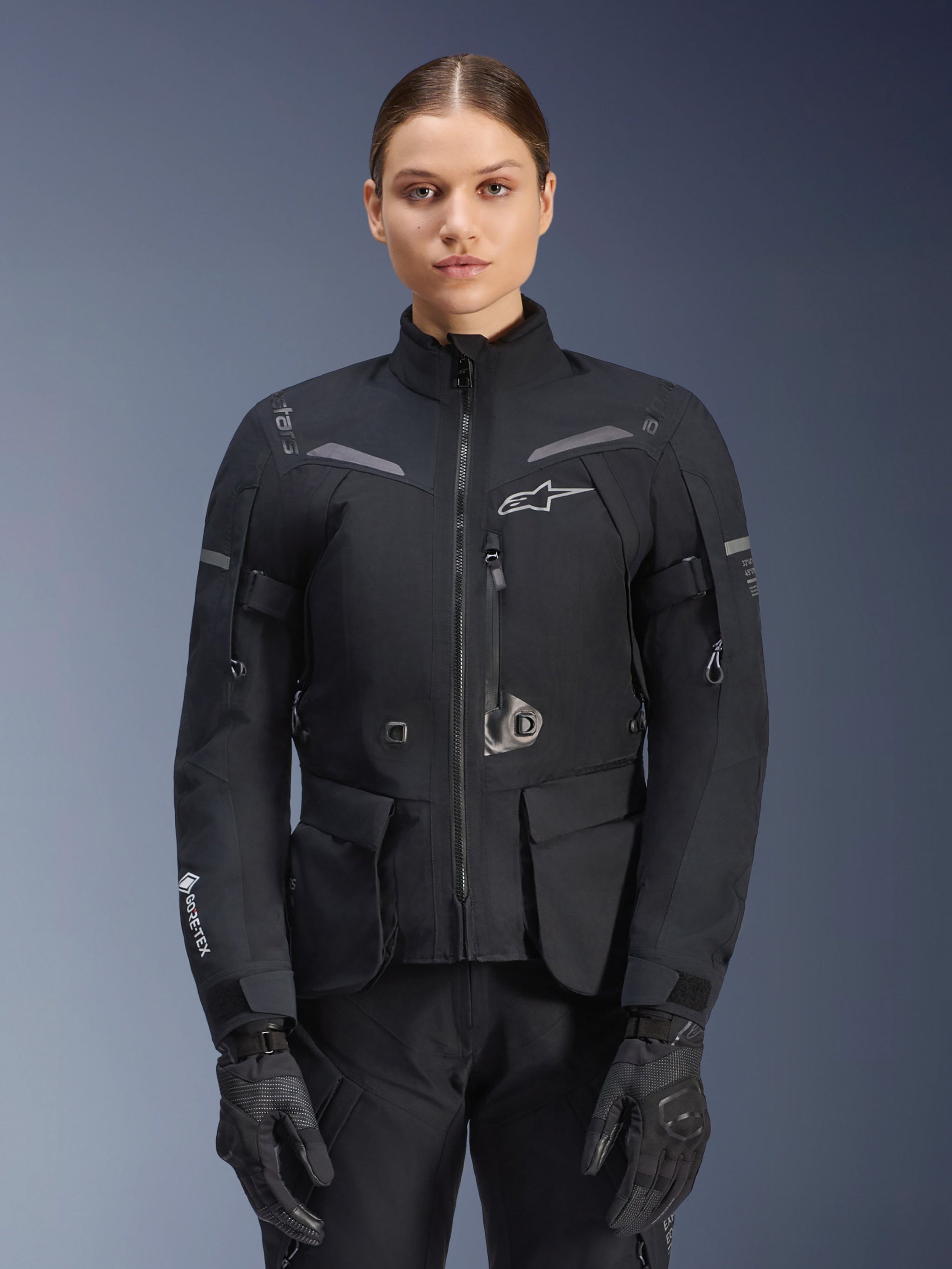 Woman wearing Alpinestars Stella ST-7 2L GORE-TEX Jacket, Black with Dark Gray accents, motorcycle touring jacket with large cargo pockets and high collar, paired with black gloves and pants, waterproof adventure riding apparel