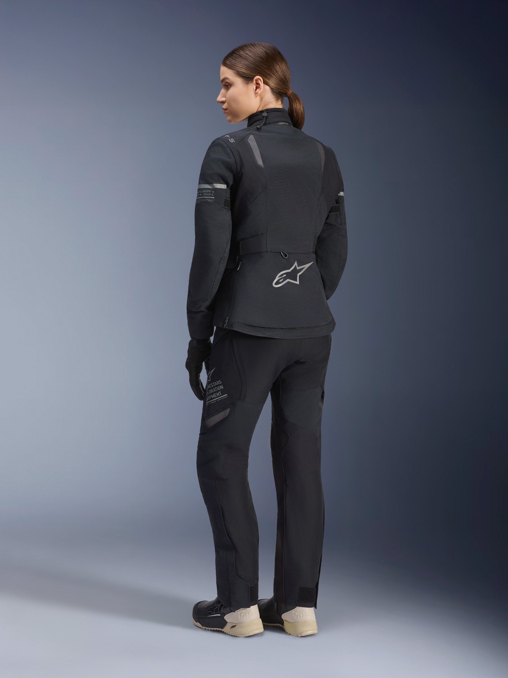 Alpinestars Women's Stella ST-7 2L GORE-TEX Jacket and Pants, full black adventure touring suit, back view showing Alpinestars star logo and Exploration Equipment branding, paired with beige boots and black gloves.