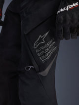 Close-up of Alpinestars ST-7 2L GORE-TEX Pants in Black, detailed view of the large thigh cargo pocket featuring gray Alpinestars Exploration Equipment branding, star logo, and geographic coordinates, high-performance textile construction for adventure touring.