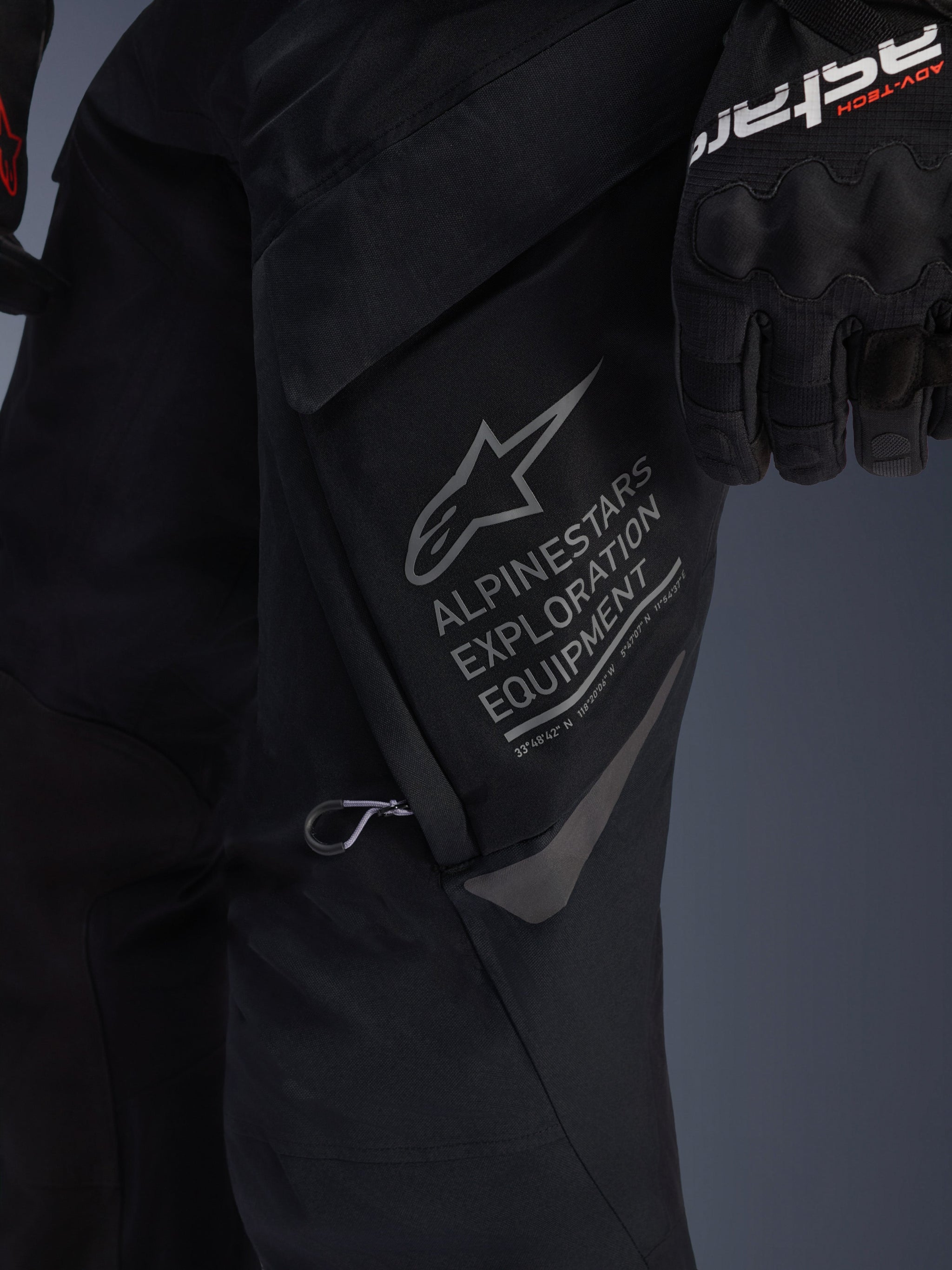 Close-up of Alpinestars ST-7 2L GORE-TEX Pants in Black, detailed view of the large thigh cargo pocket featuring gray Alpinestars Exploration Equipment branding, star logo, and geographic coordinates, high-performance textile construction for adventure touring.