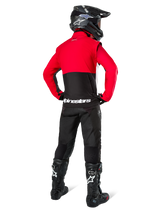 Motocross rider seen from behind wearing the Alpinestars Pro-Dura Jacket in Black, Bright Red, and White, paired with black pants, black helmet, and black off-road boots, featuring a red upper and black lower section with white Alpinestars branding across the back.