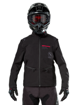 Motocross rider wearing Alpinestars Lite-Dura Softshell Jacket in Black, featuring red and grey logo details on the chest, black helmet with red-tinted goggles, black gloves and pants, front view of technical off-road riding apparel