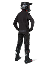 Enduro rider viewed from the back wearing the Alpinestars Lite-Dura Softshell Jacket in Black, paired with black technical riding pants and boots, featuring a zippered rear storage pocket and Alpinestars logo on the lower hem, complete off-road gear for motocross.
