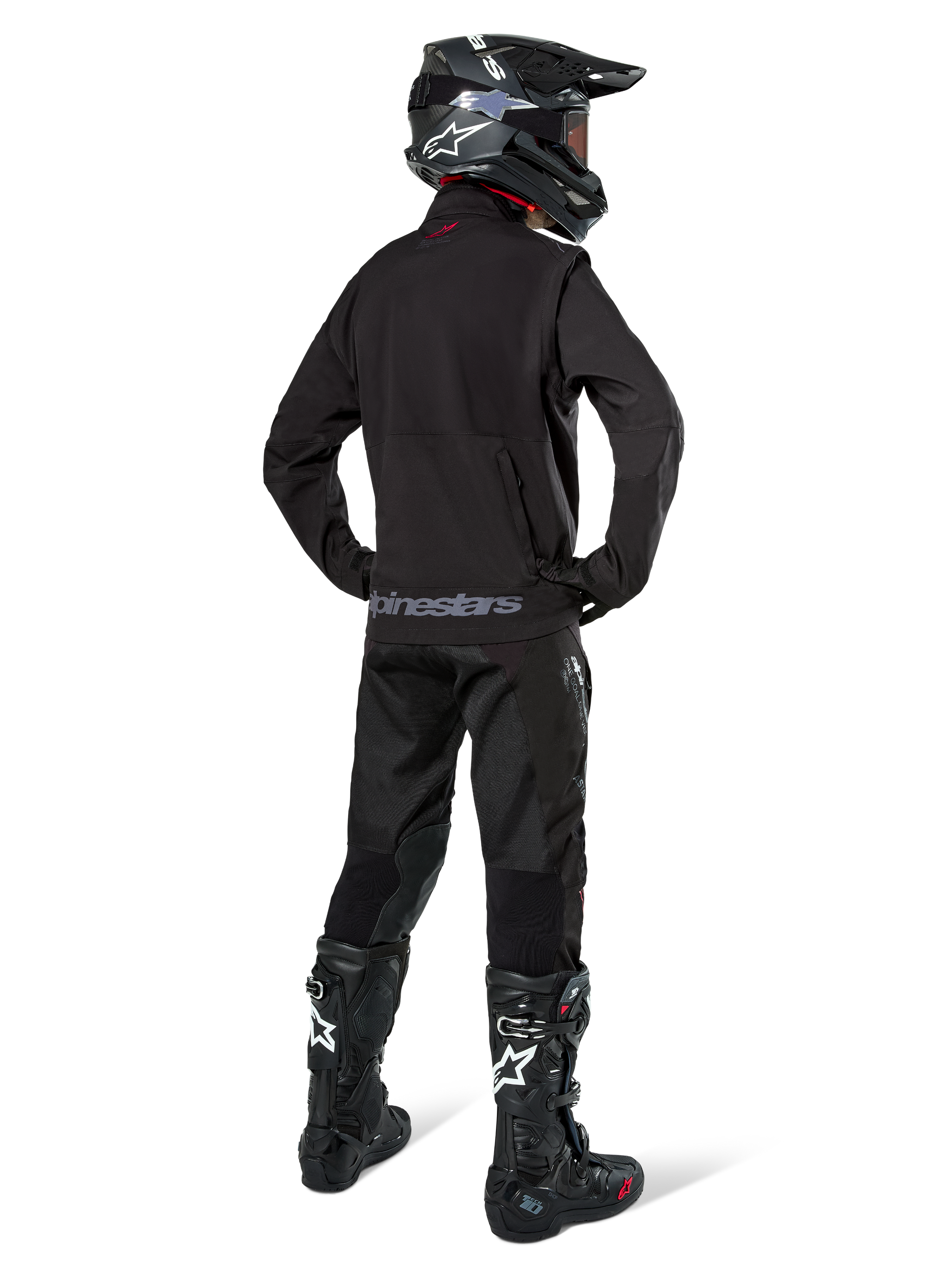 Enduro rider viewed from the back wearing the Alpinestars Lite-Dura Softshell Jacket in Black, paired with black technical riding pants and boots, featuring a zippered rear storage pocket and Alpinestars logo on the lower hem, complete off-road gear for motocross.