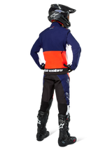 Motocross rider from the back wearing Alpinestars Lite-Dura Softshell Jacket in Navy, Hot Orange, and White, showing the large rear storage pocket. Styled with matching enduro pants, a black motocross helmet, and black off-road boots.