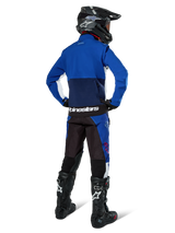 Motocross rider from behind wearing Alpinestars Lite-Dura Softshell Jacket in Navy Bright Blue and White, paired with matching enduro pants, black helmet, and black boots, complete off-road racing gear