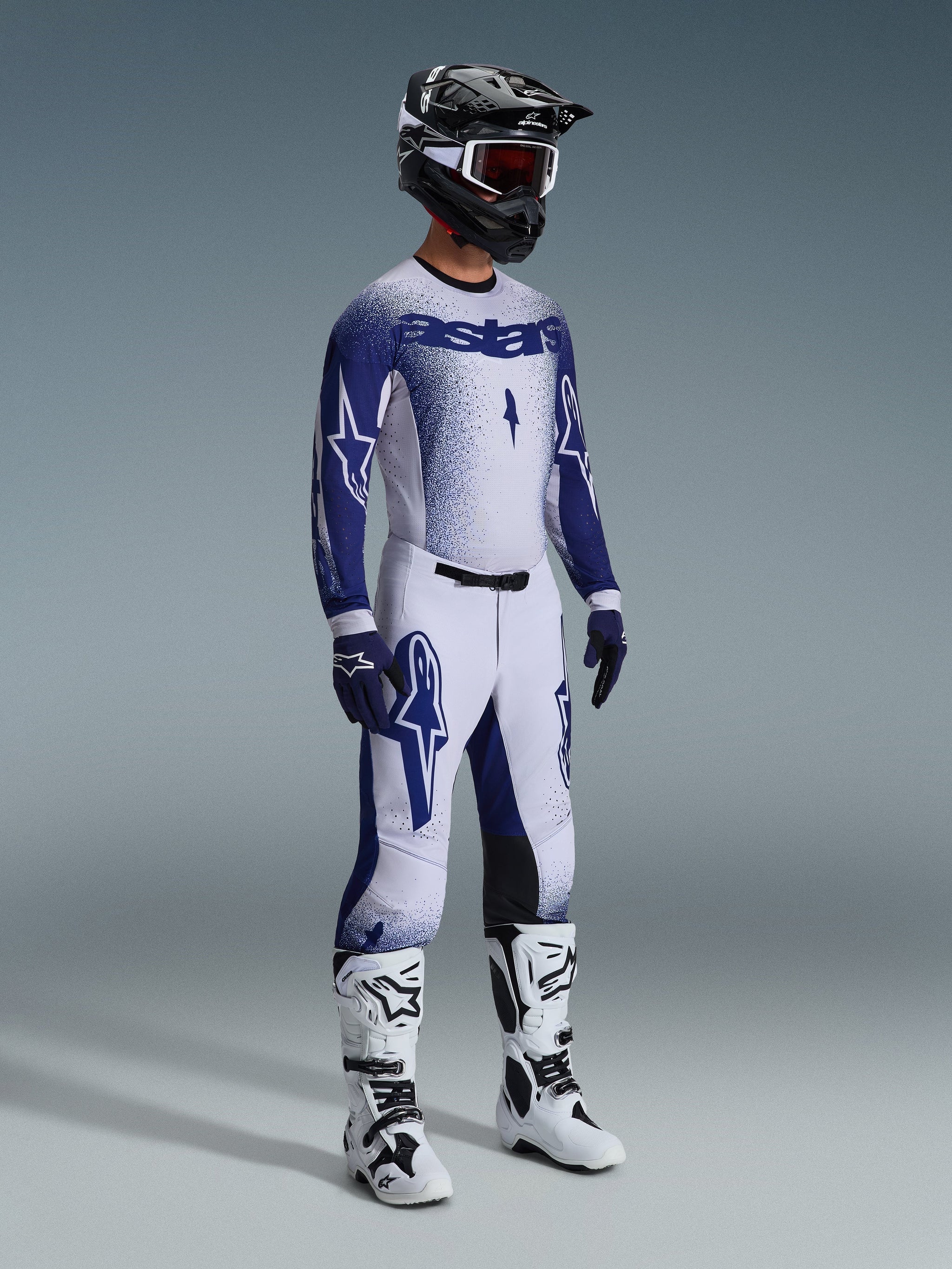 Motocross rider wearing 2026 Supertech Scenz Pants in Light Gray and Dark Blue, matching blue and white jersey with 'astars' branding and spray-paint effect, black helmet with white goggles, white and black motocross boots, coordinated professional off-road racing outfit.