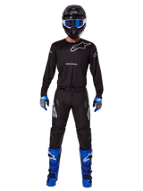 Alpinestars 2025 Racer Graphite Pants, Motocross Pants, Black and Gray, worn on a model with a matching black jersey, blue and purple boots, and blue goggles, complete off-road racing outfit featuring bold Alpinestars branding