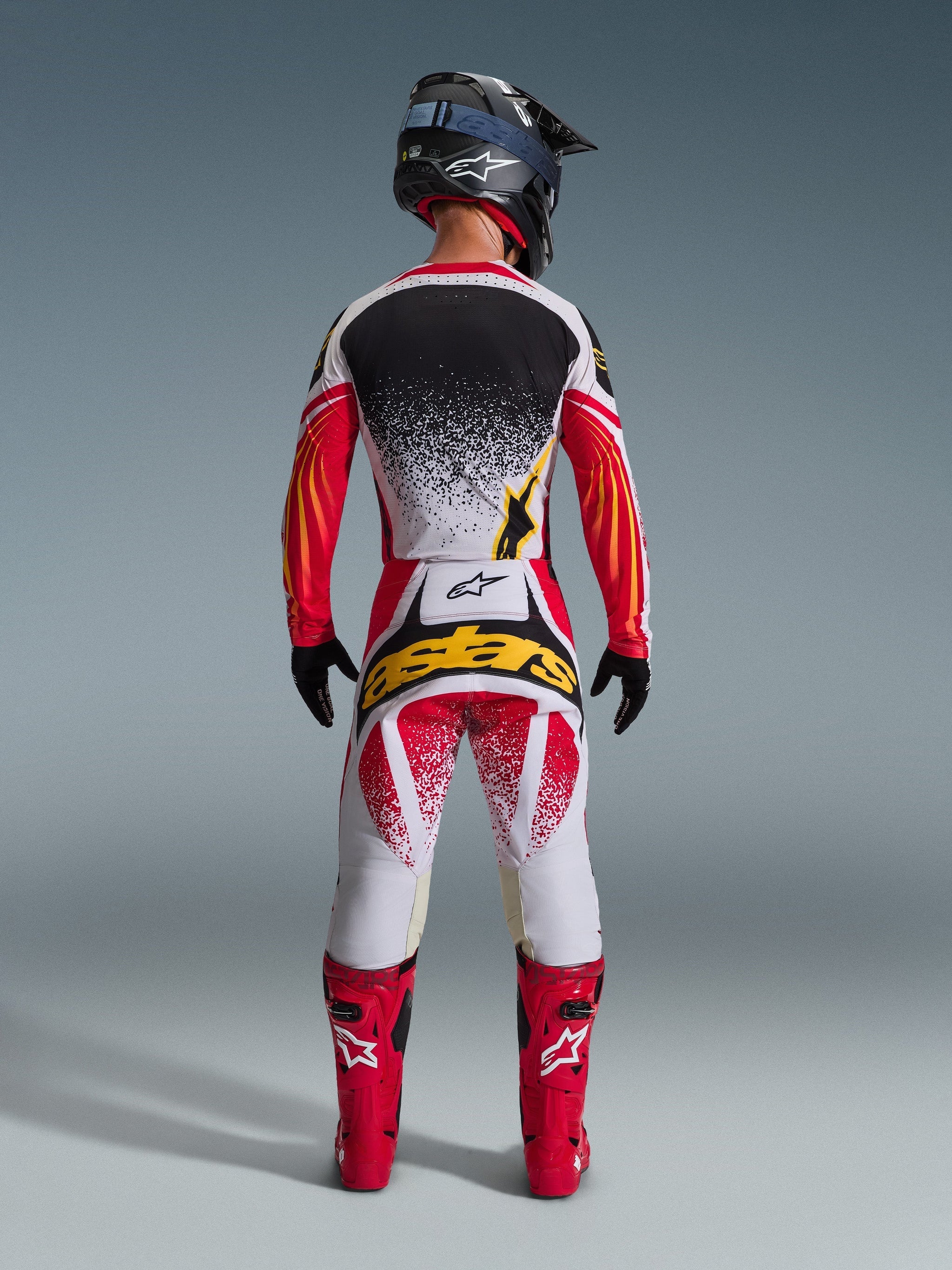 Motocross rider from behind wearing complete racing gear, black and white jersey with red sleeves and yellow accents, 2026 Techstar Nomur Pants in white and red with a large yellow 'astars' logo on the back, bright red boots, and black helmet, coordinated outfit for off-road racing