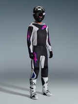 Motocross rider wearing complete racing gear, Alpinestars 2026 Fluid Grid Pants in Black Light Gray and Purple, matching jersey with pink and blue graphics, black helmet and goggles, white and black boots, and black gloves, coordinated outfit for off-road racing