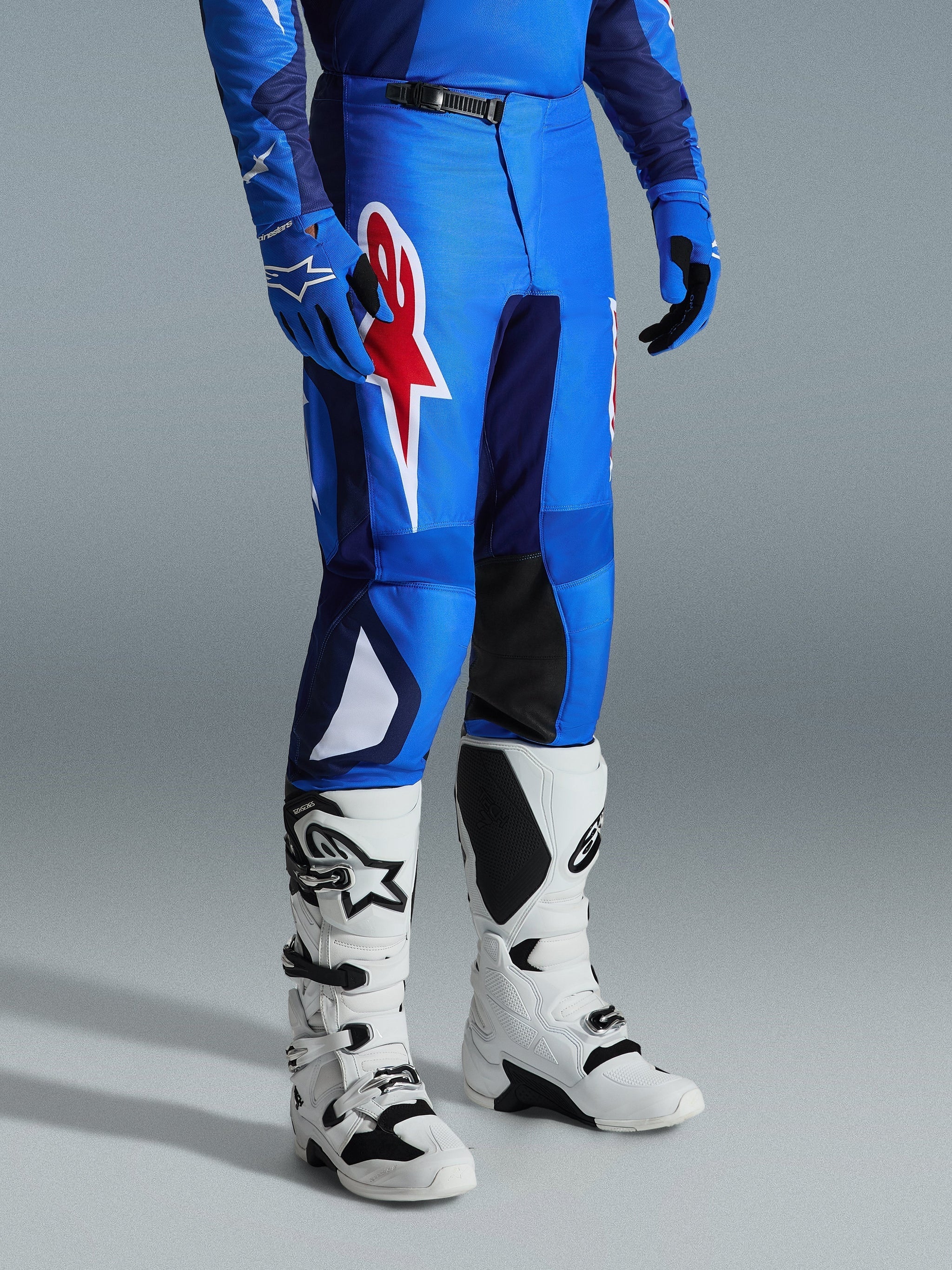 Alpinestars 2026 Fluid Grid Pants, Motocross Pants, Blue and Red, worn with white motocross boots and blue gloves, featuring a large red star logo on the thigh and black inner knee panels, designed for off-road riding.