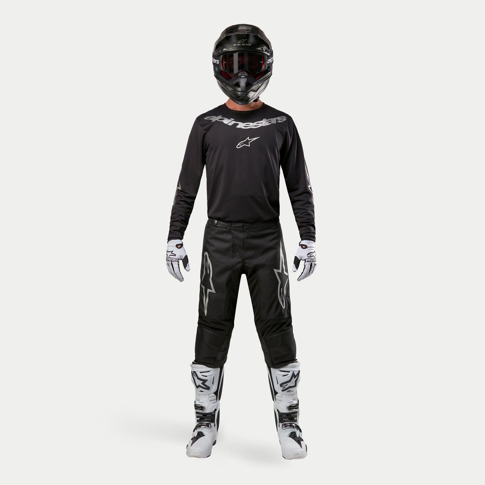 Motocross rider wearing complete racing gear, Alpinestars 2024 Fluid Graphite Pants in Black and Silver, black long-sleeve jersey with silver branding, black helmet with goggles, white and black boots, white and black gloves, coordinated outfit for motocross