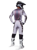 Motocross rider wearing complete racing gear, grey and black jersey, light grey pants with black 'Astars' branding on the sides, black and white helmet, White Tech 10 Boots, coordinated professional off-road racing outfit