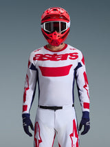 Motocross rider wearing the Alpinestars 2026 Racer Riway Jersey in White Blue Red, featuring a large red 'astars' logo on the chest, navy blue side panels, matching white and red motocross pants, a red helmet with white goggles, and navy blue gloves, complete off-road racing gear set.